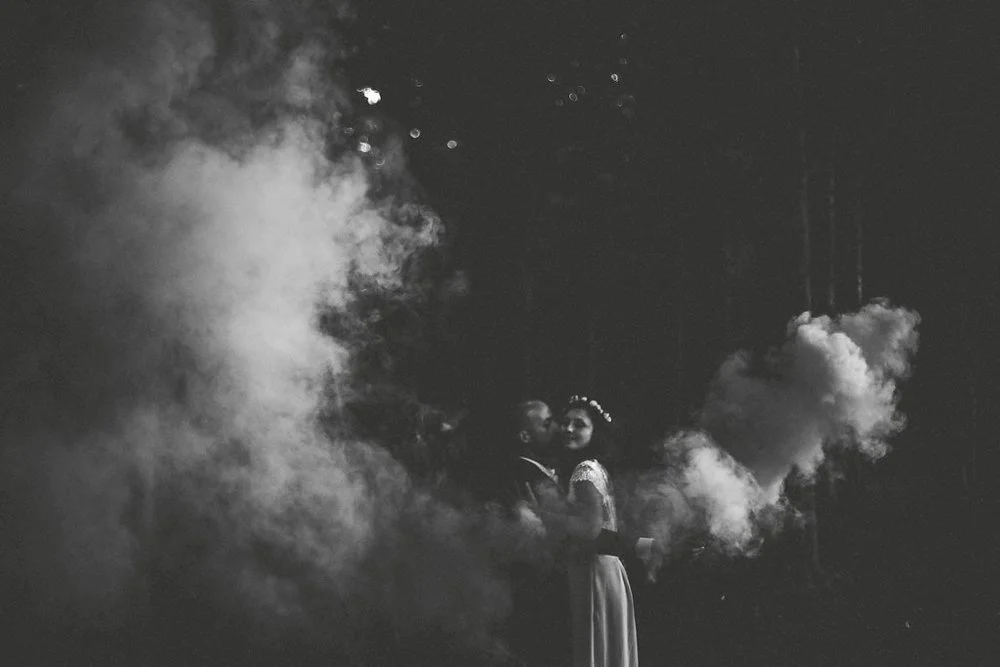 Un couple danse dans un champ de fumée ou de vapeur avec un fond sombre, en noir et blanc.