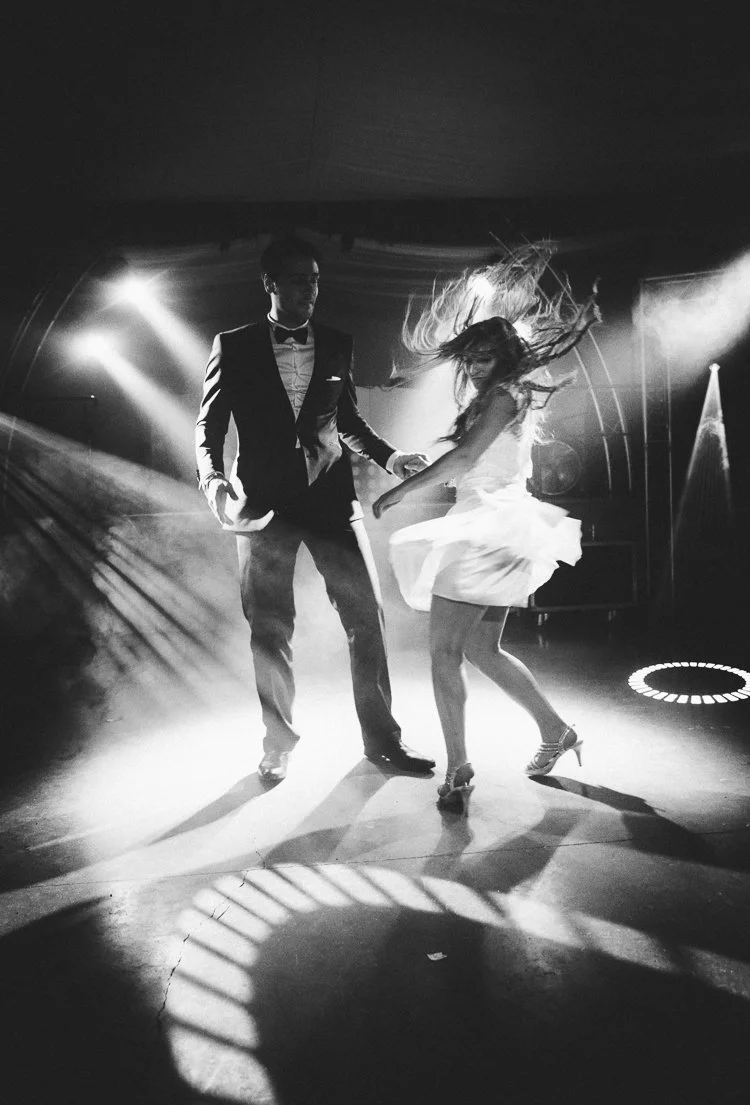 Un homme et une femme dansent sur une scène éclairée, la femme tourne avec une robe blanche en mouvement. La scène est sombre avec des jeux de lumières et d'effets de lumière. C'est une scène de danse ou un spectacle.