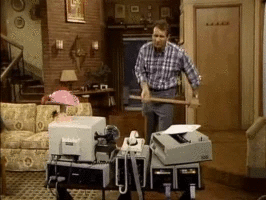 Un homme berçant une fidèle dans un salon, entouré d'équipements de bureau et de machines, dans une scène de sitcom.