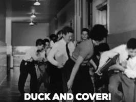 Groupe de personnes jouant à un jeu de canard et de couverture dans un couloir, avec texte "DUCK AND COVER" en bas.