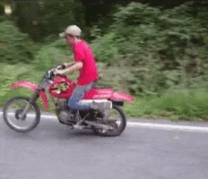 Personne portant un casque rouge, en t-shirt rose, roulant à moto sur une route.