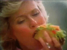 Une femme mange un sandwich ou une grande tartine avec divers ingrédients, dans un contexte intérieur.