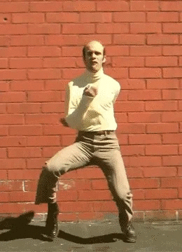 Une personne danse en face d'un mur en briques rouges, avec un pantalon gris et un haut beige, dans une pose dynamique.