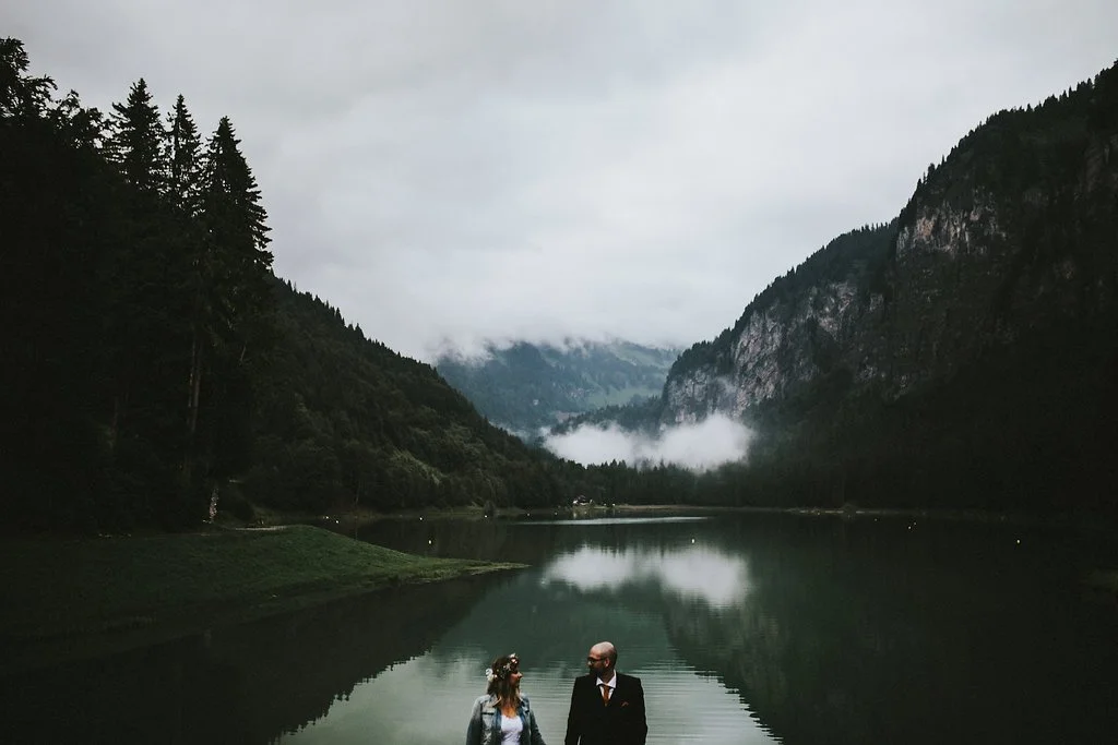 Un couple habillé élégamment marche près d'un lac dans une vallée entourée de montagnes et d'arbres, avec un ciel nuageux.