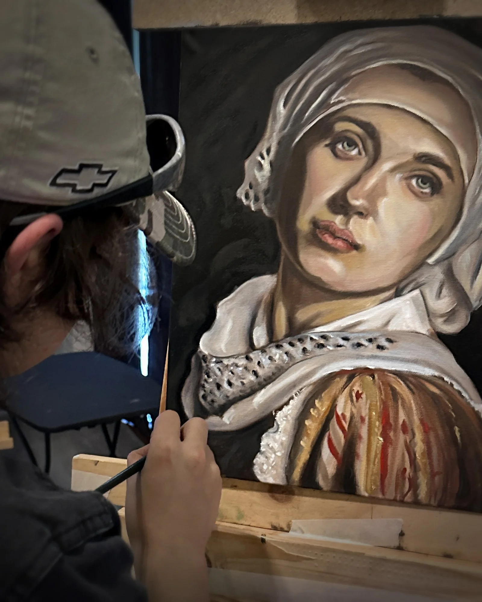 A painting of a woman with blue eyes wearing a white headscarf and a white garment adorned with black patterns, being painted with oil paints, with a painter's hand visible holding a brush, and a container of black paint nearby.