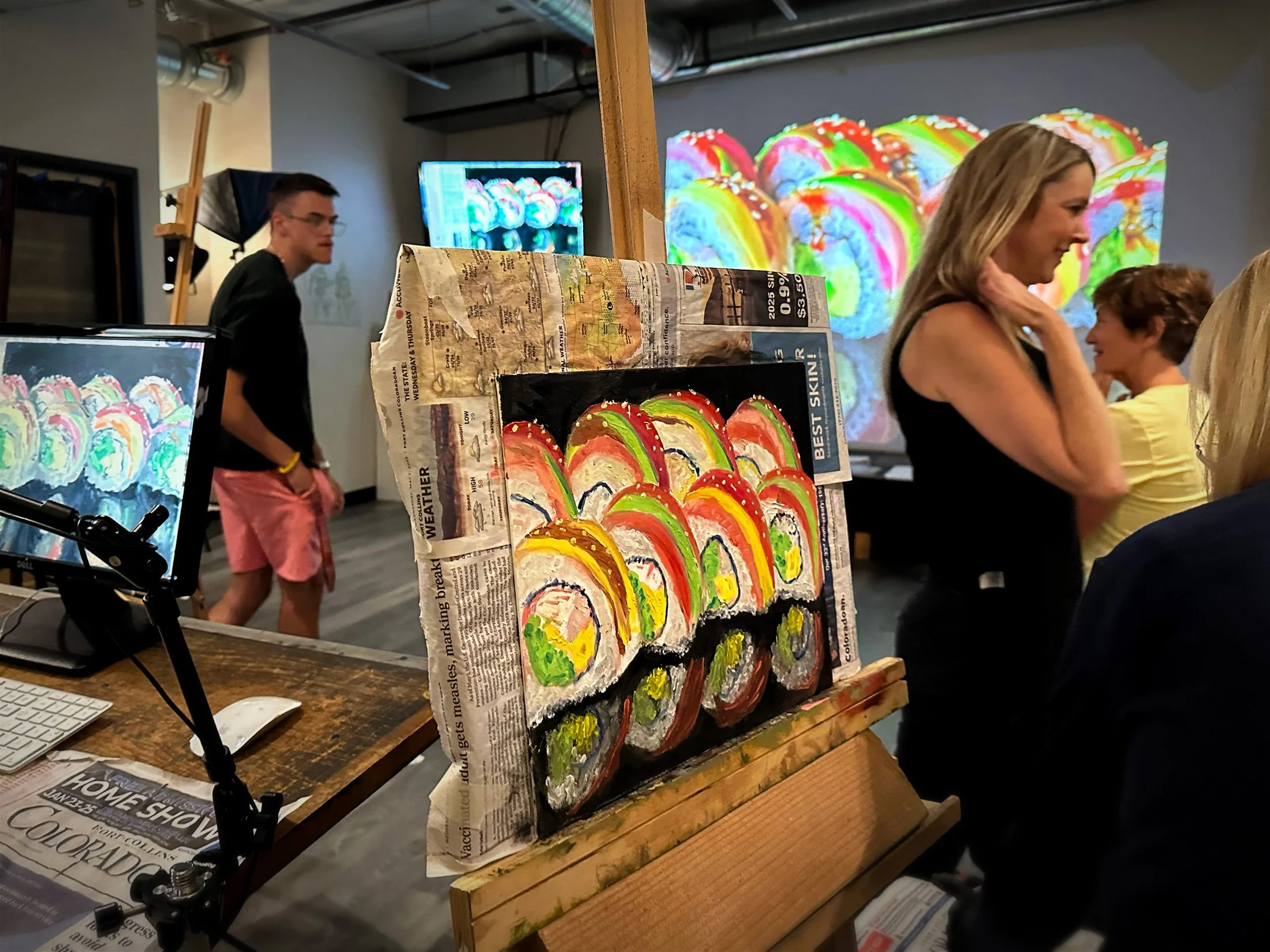 A colorful abstract painting resembling rainbow swirl patterns on a canvas, displayed on an easel with newspaper covering parts of it, in an indoor setting with several people in the background.
