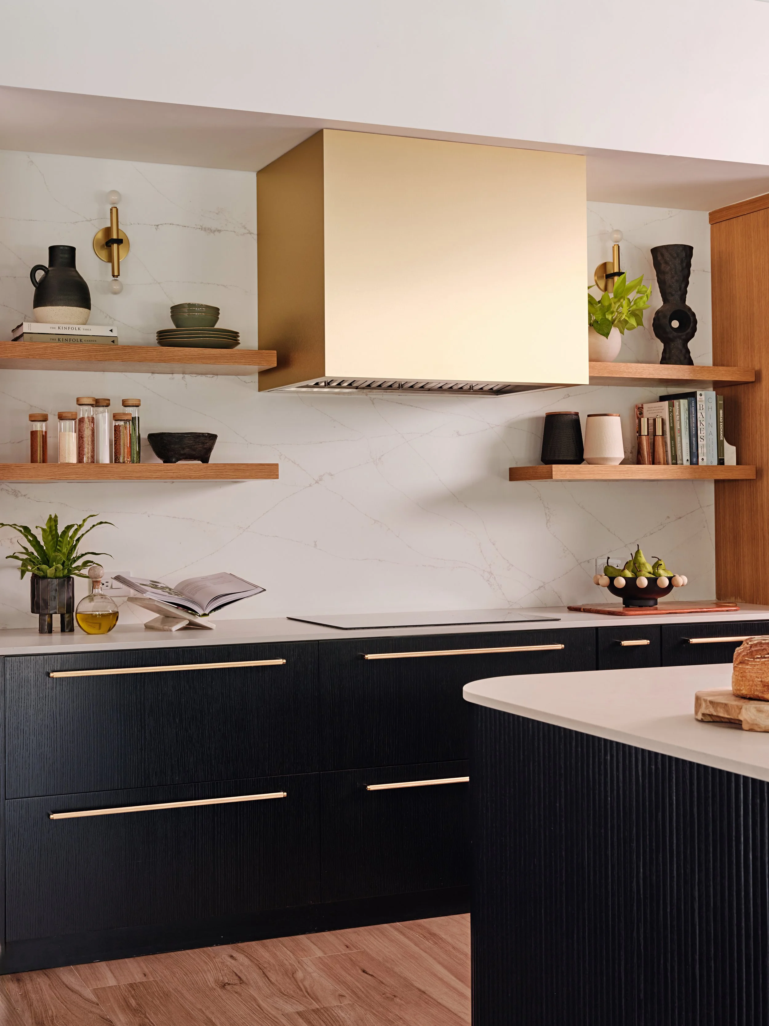 Hoodfan cladded in brass style materials. Floating oak open shelves in kitchen in Miami, FL. Interior design by Concept 2 Design.