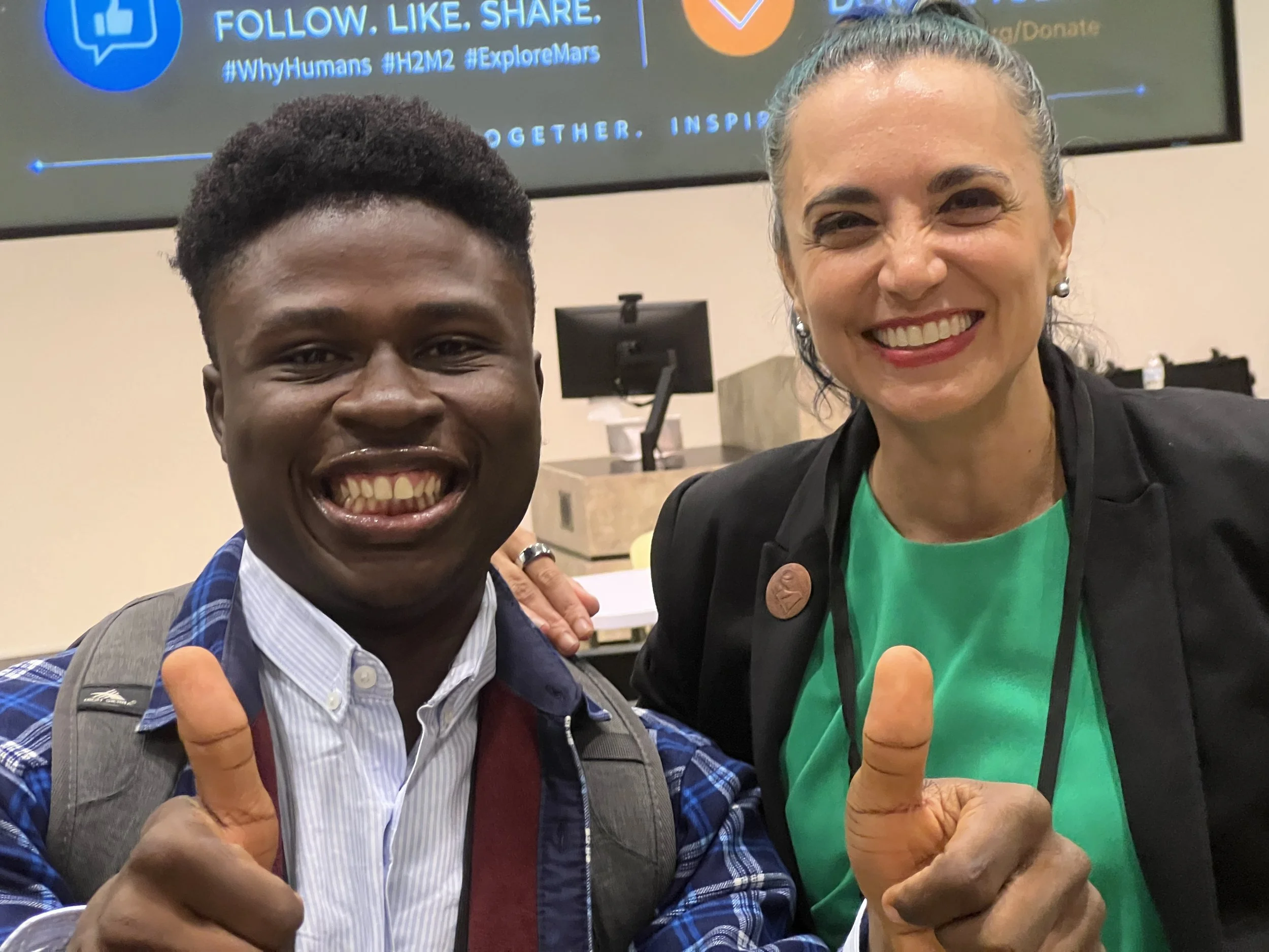 Tiffany Vora and Oluwatosin Kolade smile and give thumbs up for a photo at the 2026 Humans to the Moon and Mars Summit, with a presentation screen visible in the background.
