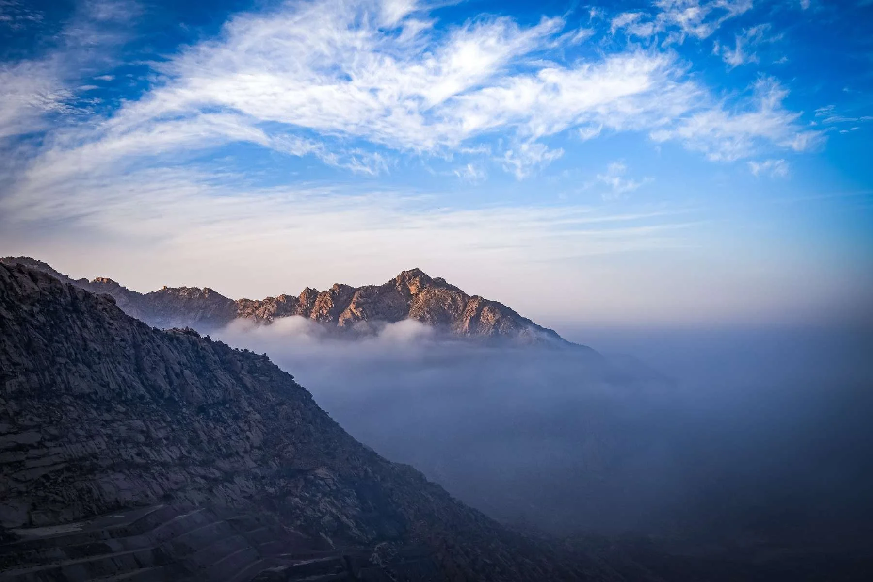 A dramatic view of rugged mountain peaks in Al Hada, Saudi Arabia, partially obscured by a thick layer of white mist and clouds under a bright blue sky.