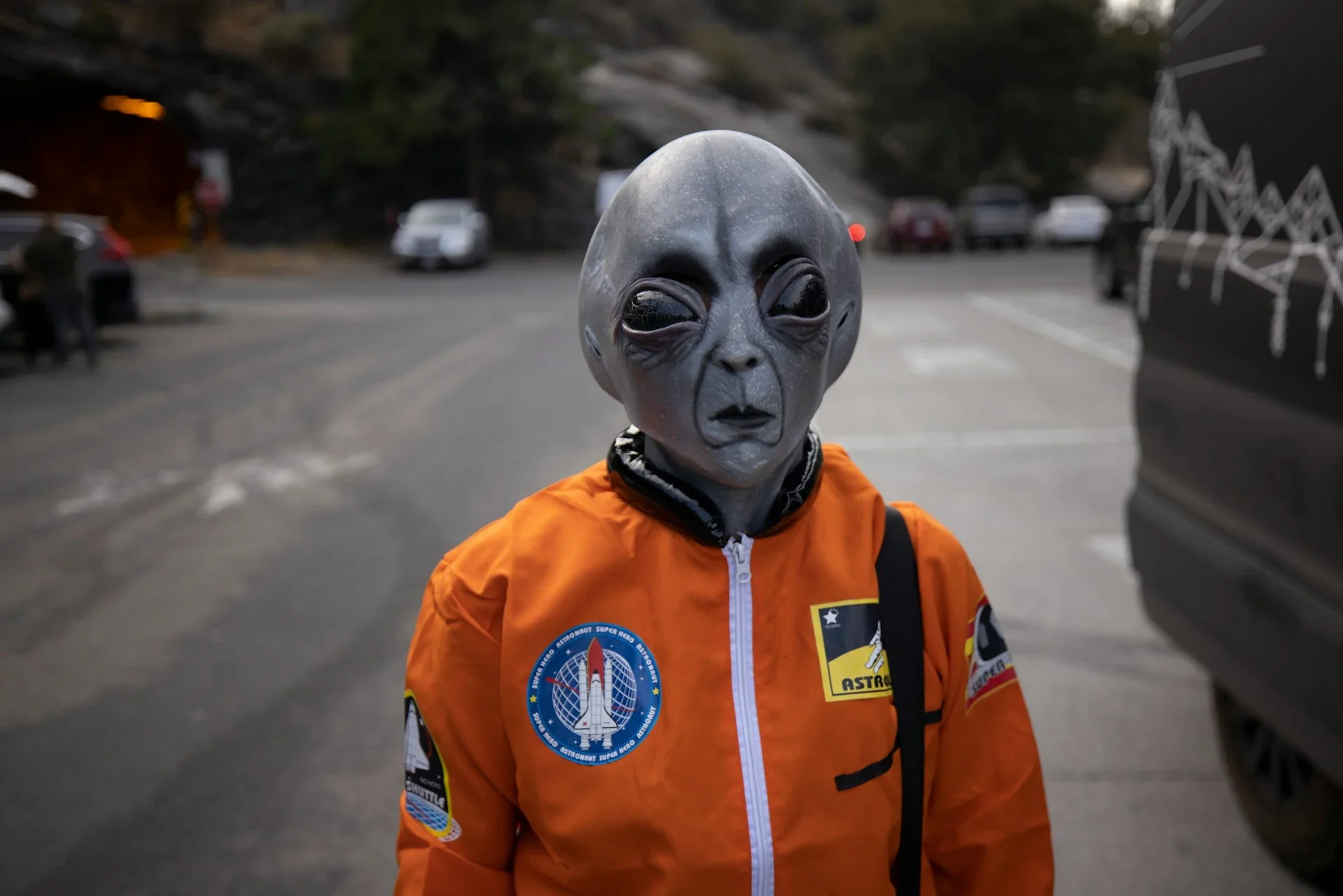 Person wearing an alien mask and orange clothing standing on a city street.