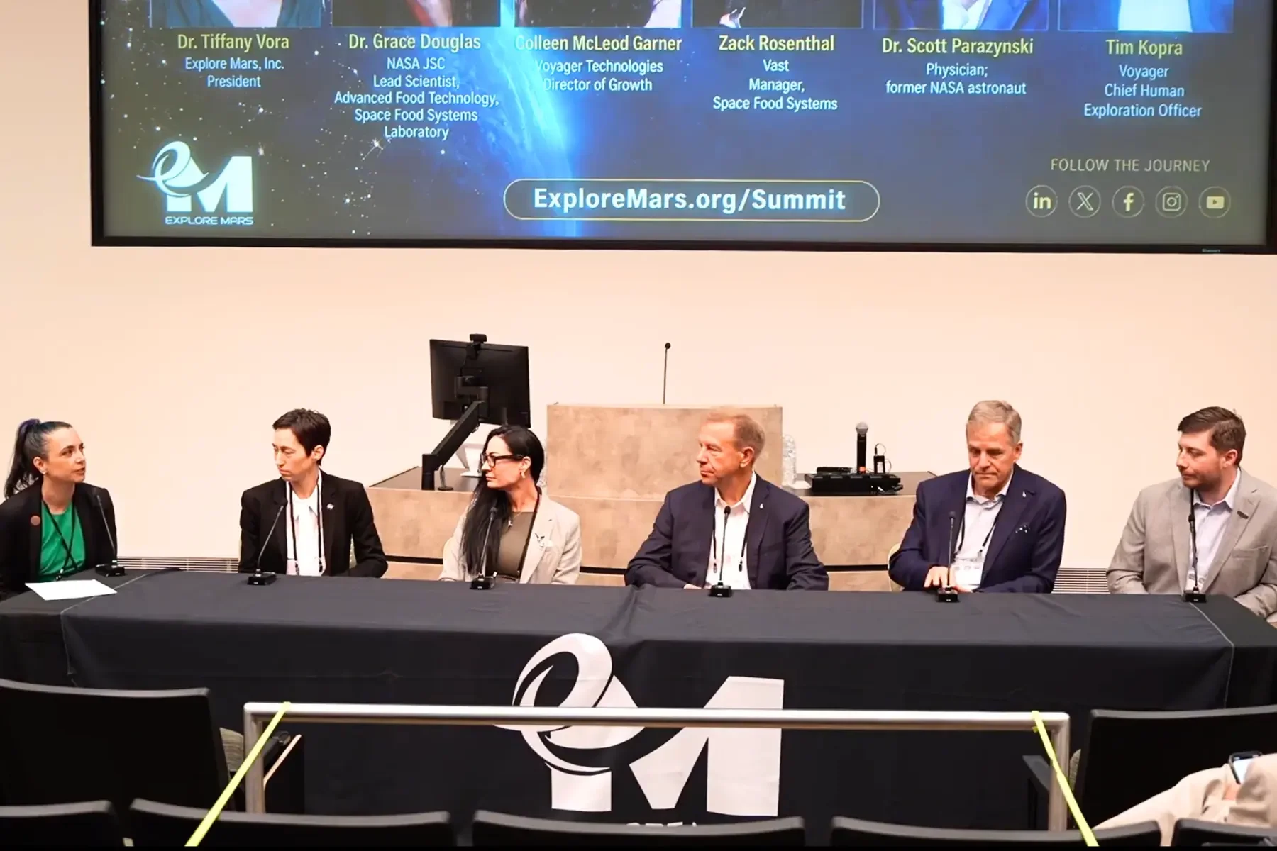 Six panelists seated at a long table on stage, each with a microphone and name placard displayed above them, at the Explore Mars Humans to the Moon and Mars Summit.