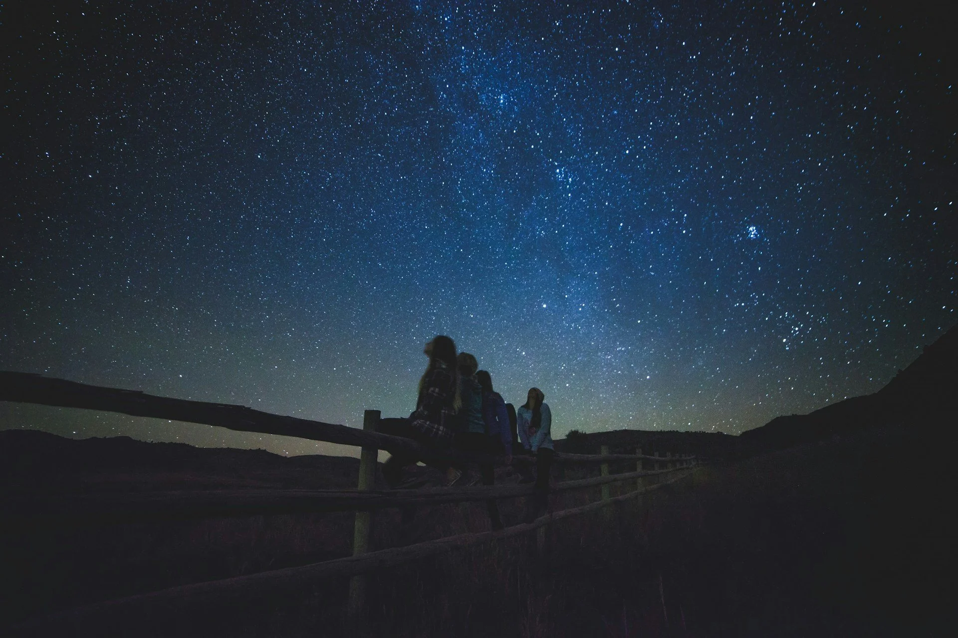 A silhouette of four people sitting on a wooden fence at night, looking up at a brilliant, star-filled sky dominated by the Milky Way galaxy.