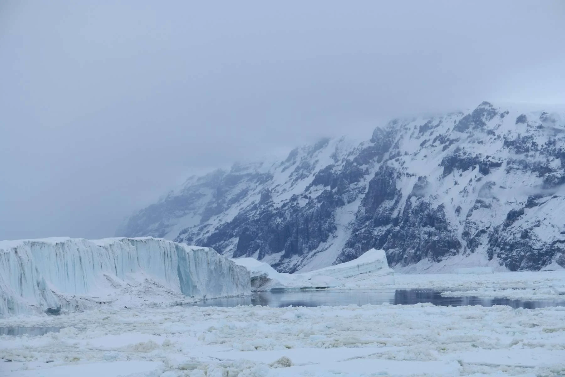 A vast, icy mountain range and glacier meet the frozen Ross Sea in Antarctica under a cloudy, atmospheric sky.