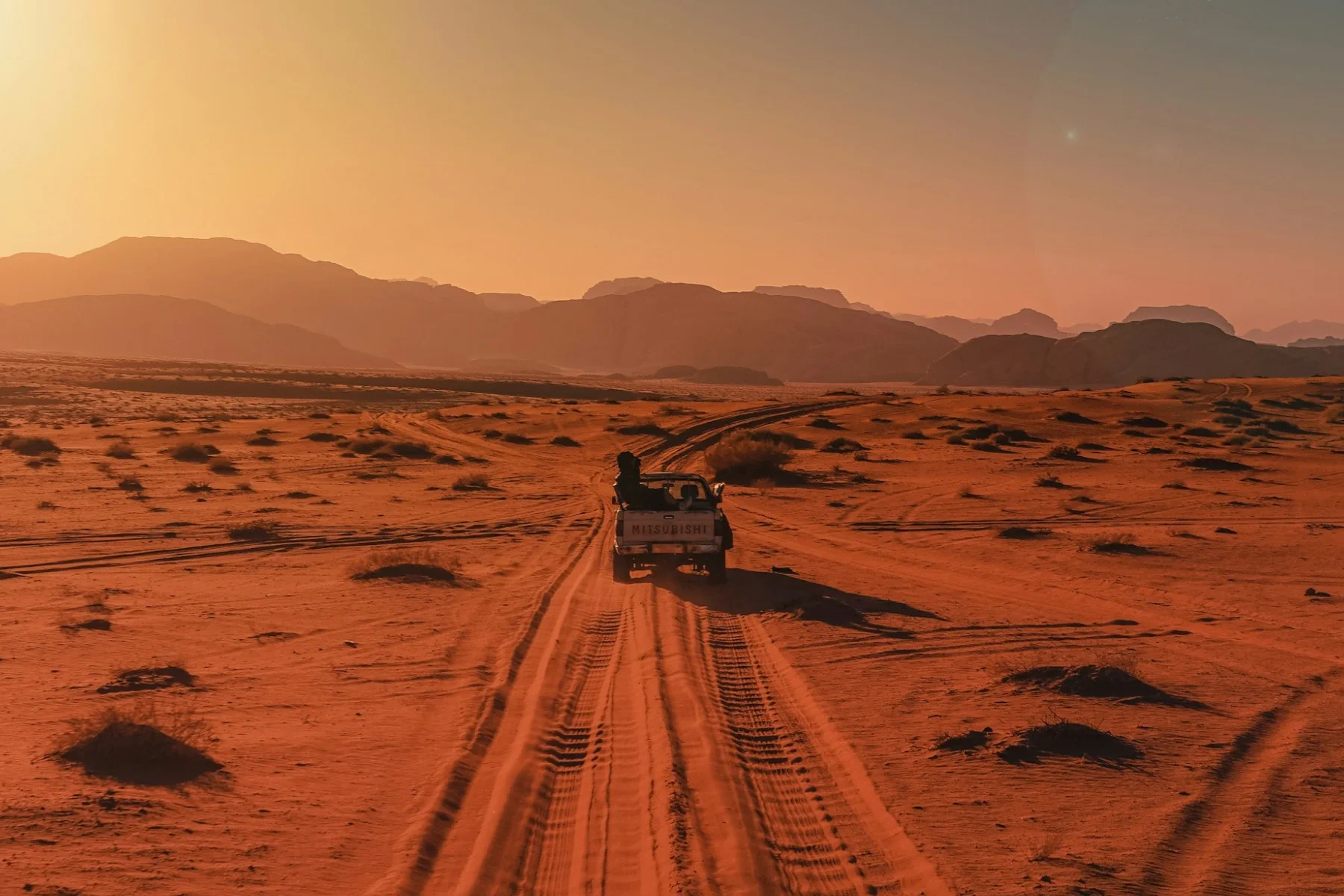 A vehicle travels along a dirt track through a vast red, rocky landscape beneath an orange sky, evoking the surface of Mars.