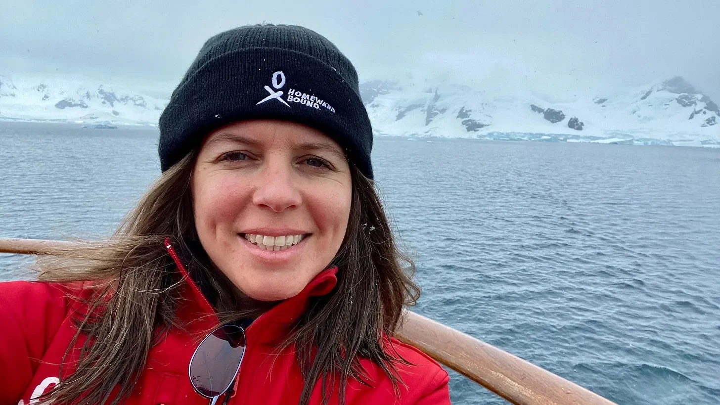 A headshot of Dr. Anna Frebel smiling directly at the camera. In the background, a calm, grey sea is visible, with distant, snow-capped mountains under a clouded Antarctic sky.