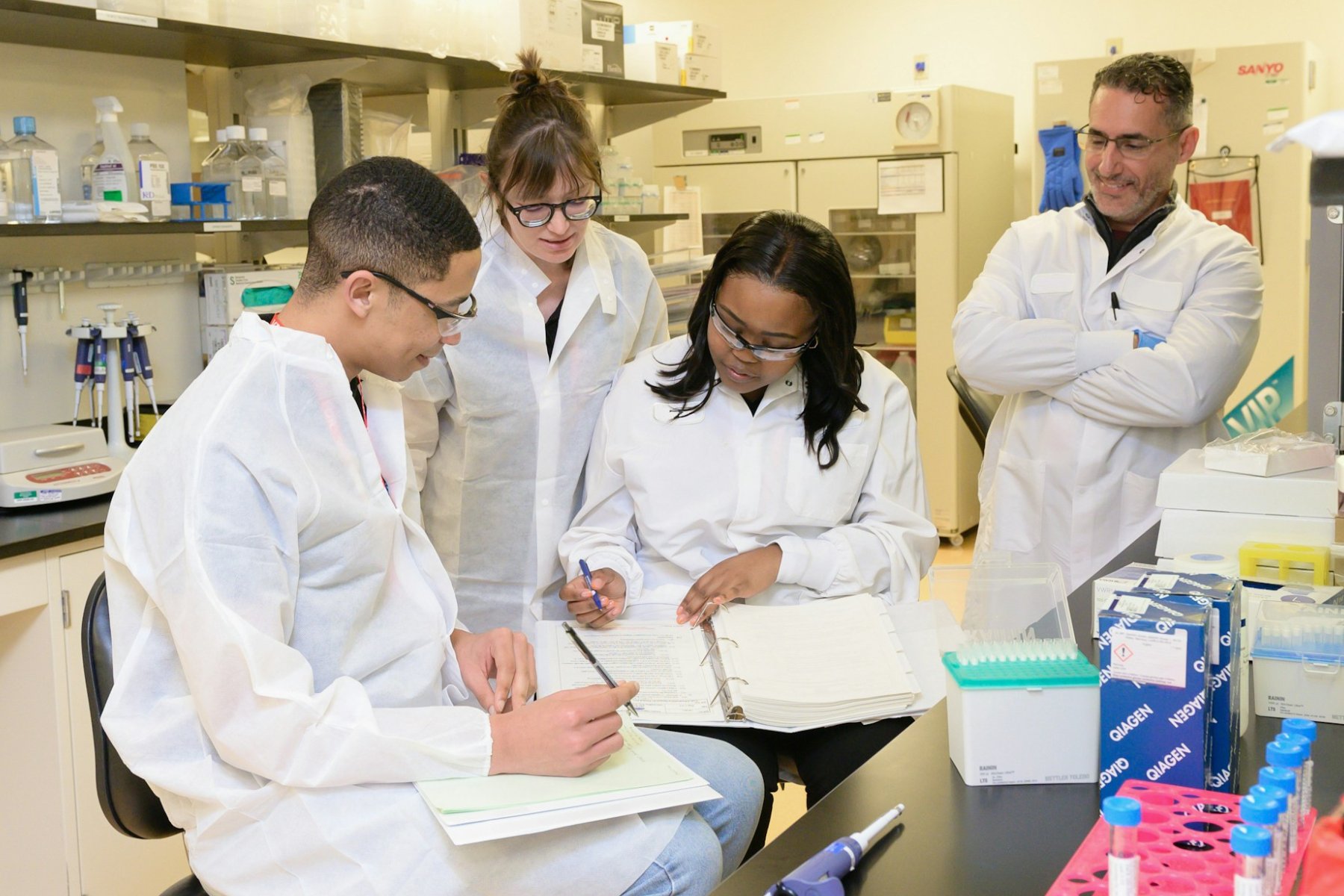 Four scientists in white lab coats and goggles working collaboratively in a professional laboratory to ensure safety and rigor.
