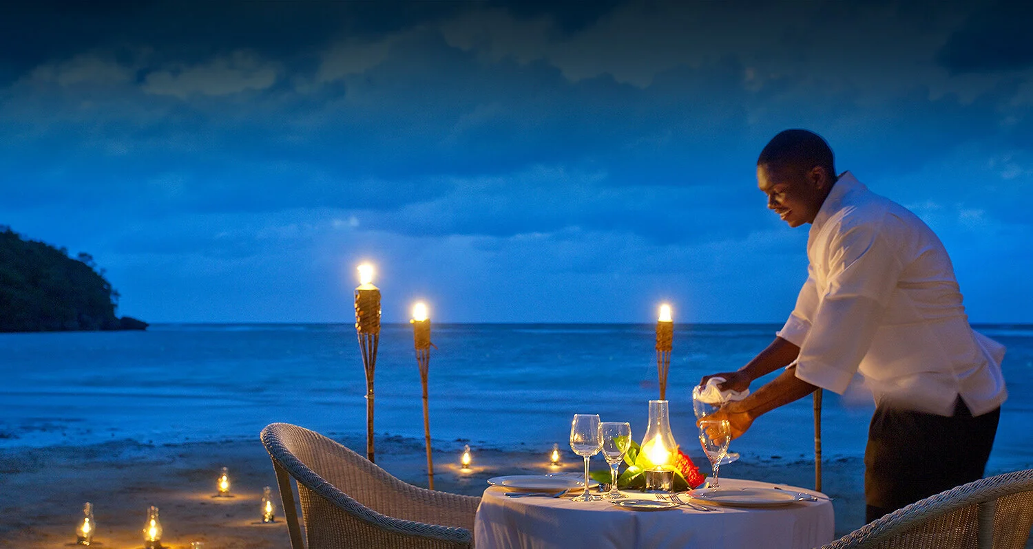 A waiter setting a romantic dinner table with wine glasses and candles on a beach at dusk, with tiki torches and the ocean in the background.