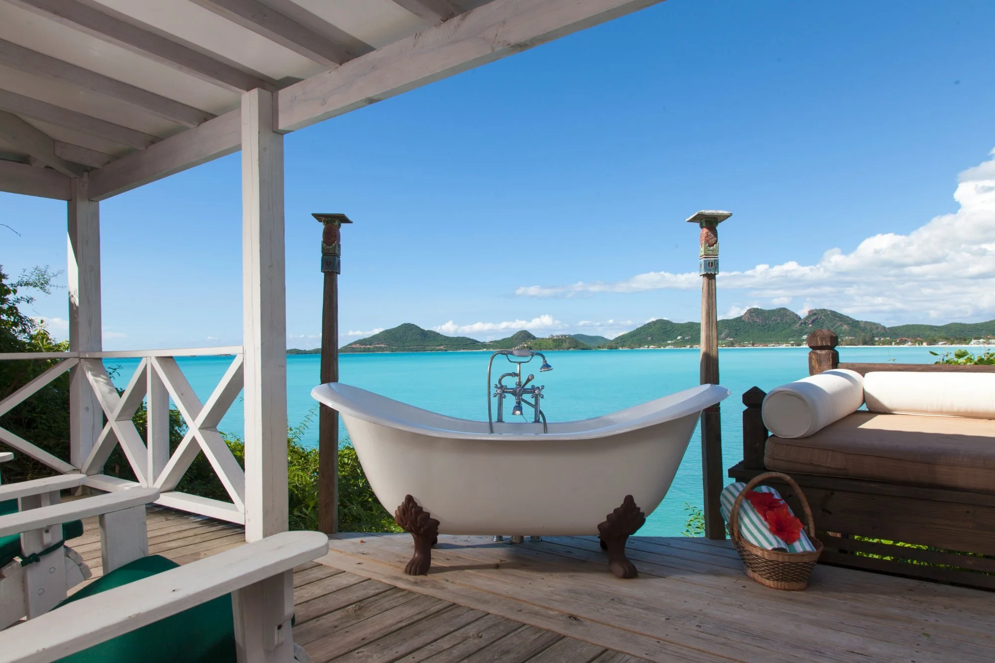 Outdoor porch with a vintage bathtub facing a turquoise body of water and distant green mountains under a partly cloudy sky.