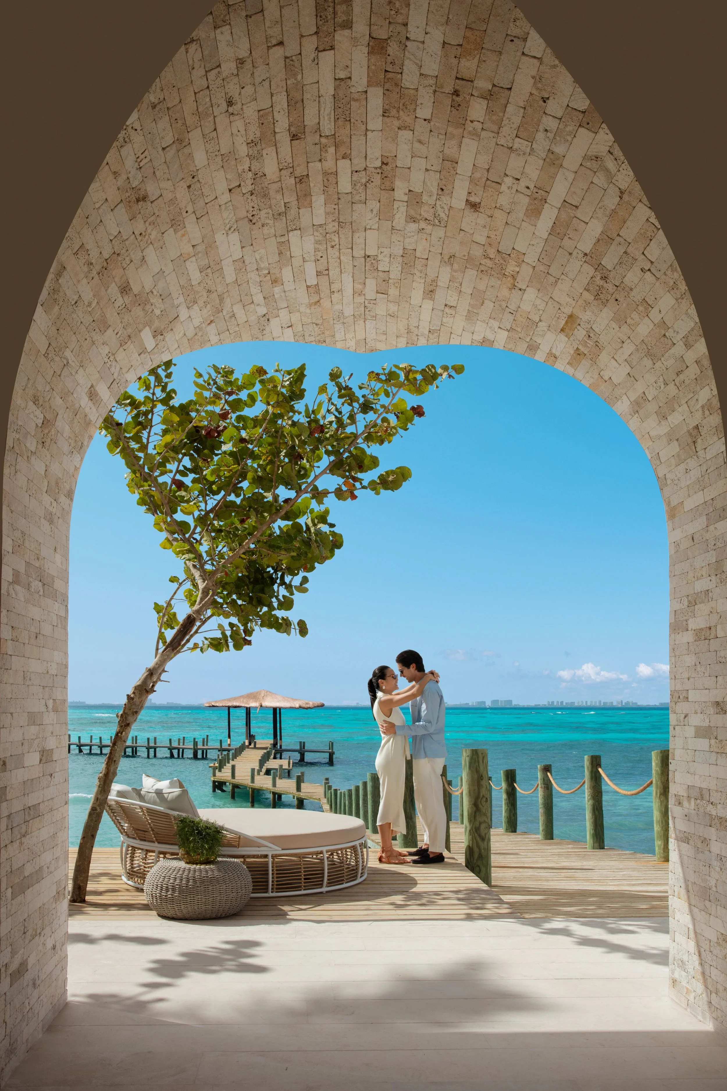 A couple embraces on a wooden dock over turquoise water, with a thatched-roof hut in the background, seen through a stone archway, under a bright blue sky.