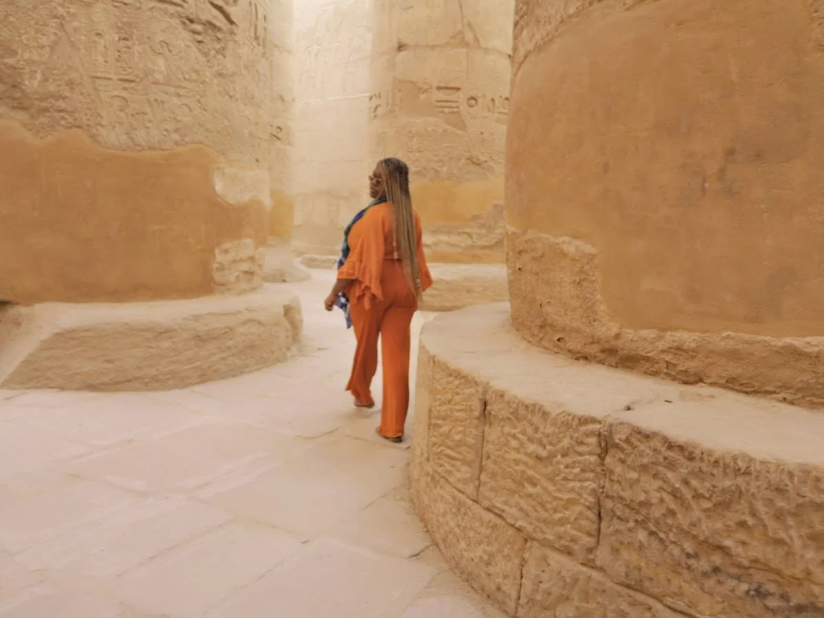 A woman dressed in orange pants and a matching top, with a blue scarf, is walking inside an ancient Egyptian temple or monument with large sandstone columns and walls.