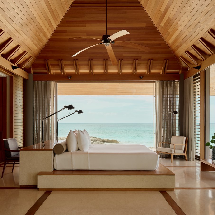 A bedroom with a wooden ceiling and wall, featuring a large bed with multiple pillows, facing a window with a view of the ocean and sky.