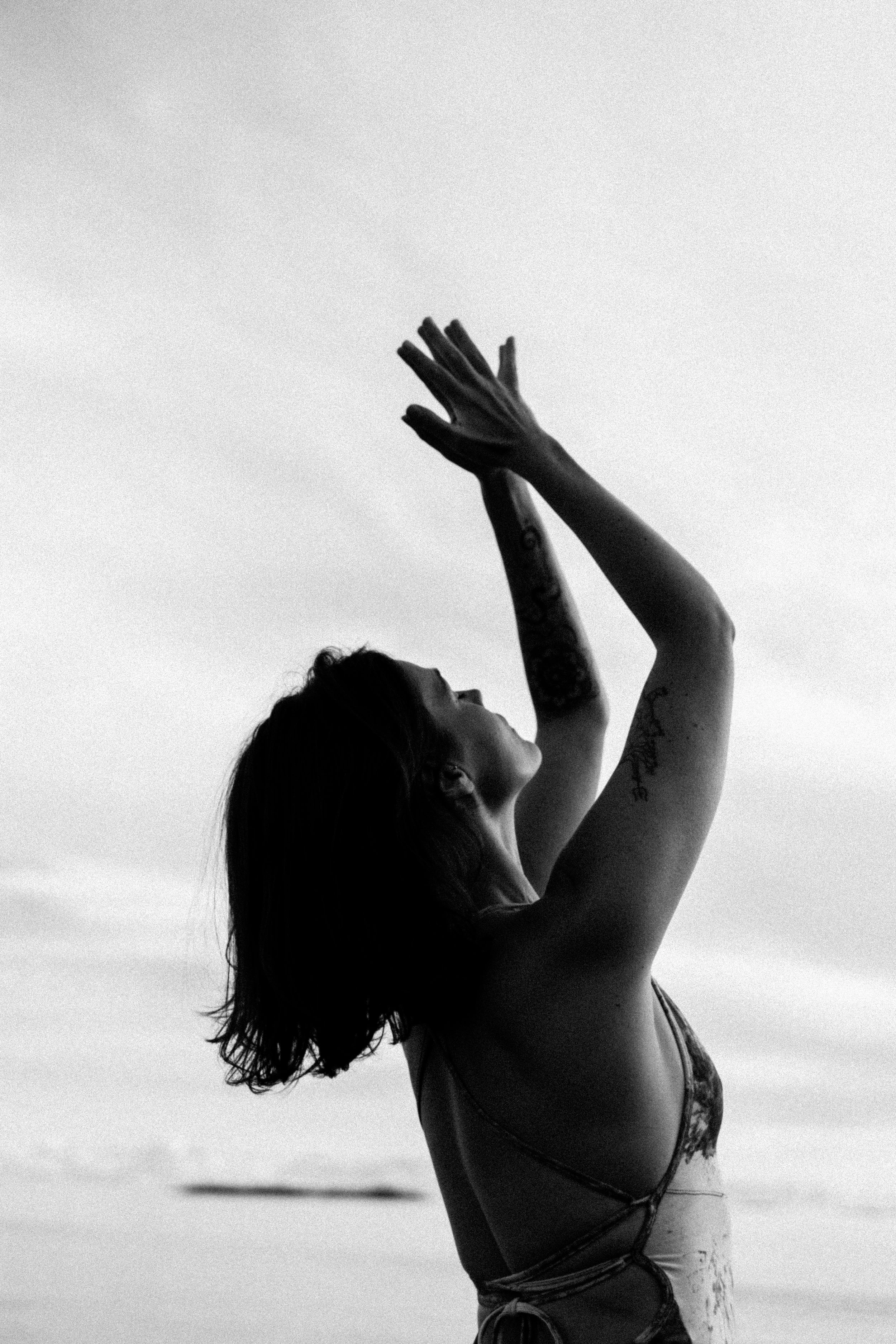 A woman with tattoos on her arm, in profile, raising her hands above her head, on a beach with a cloudy sky.