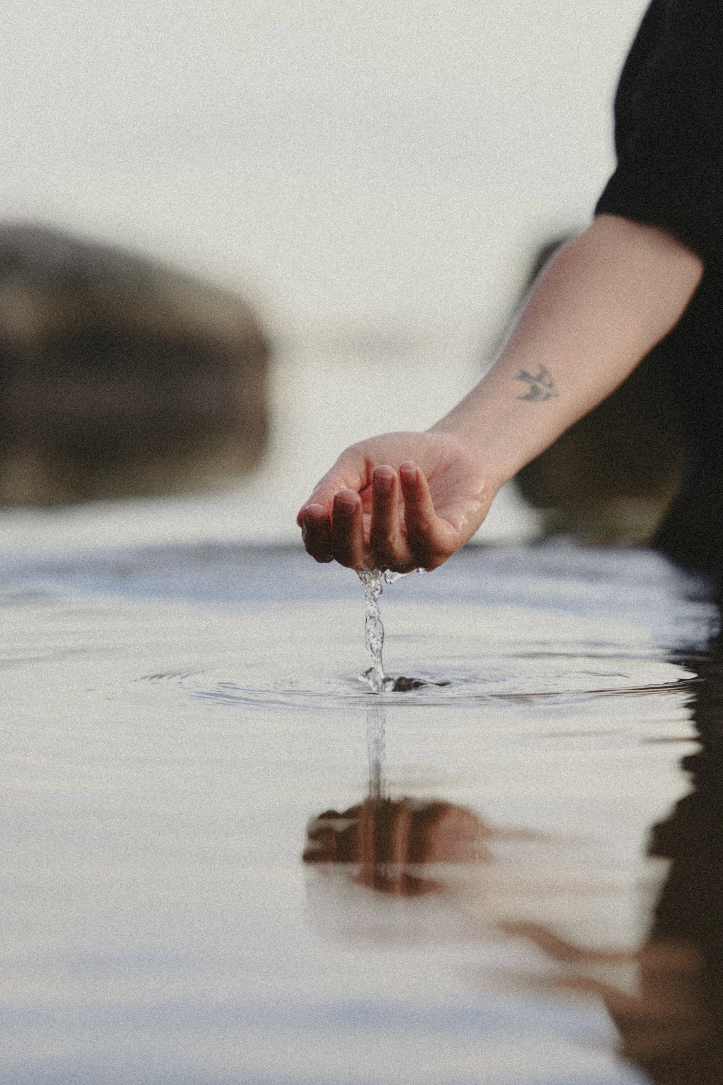 A person with a tattoo on their arm is touching water with their hand at the edge of a body of water, creating ripples.