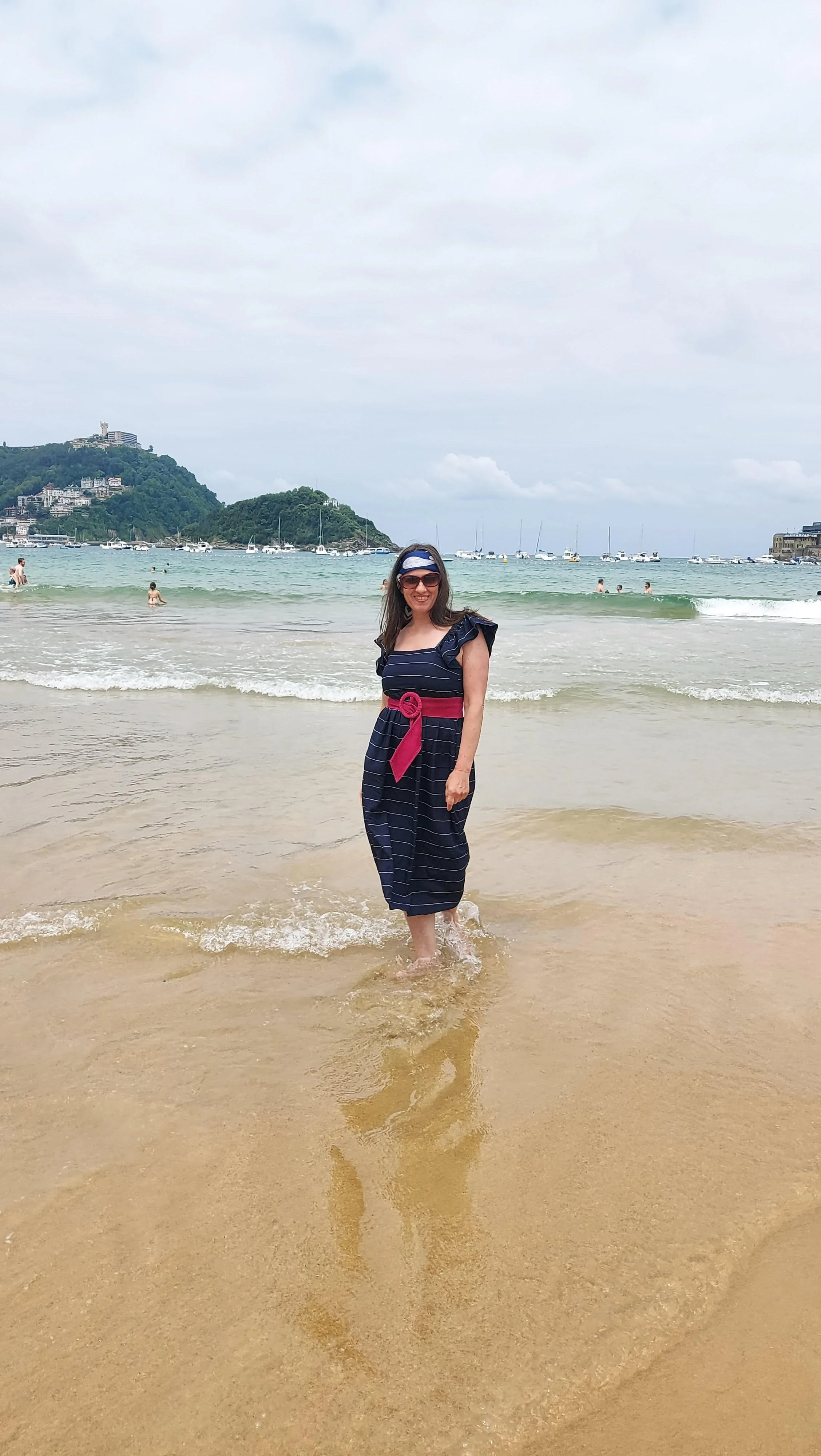 Woman in a navy blue dress with pink sash walking in the ocean at the beach, smiling, with boats and a hill in the background.