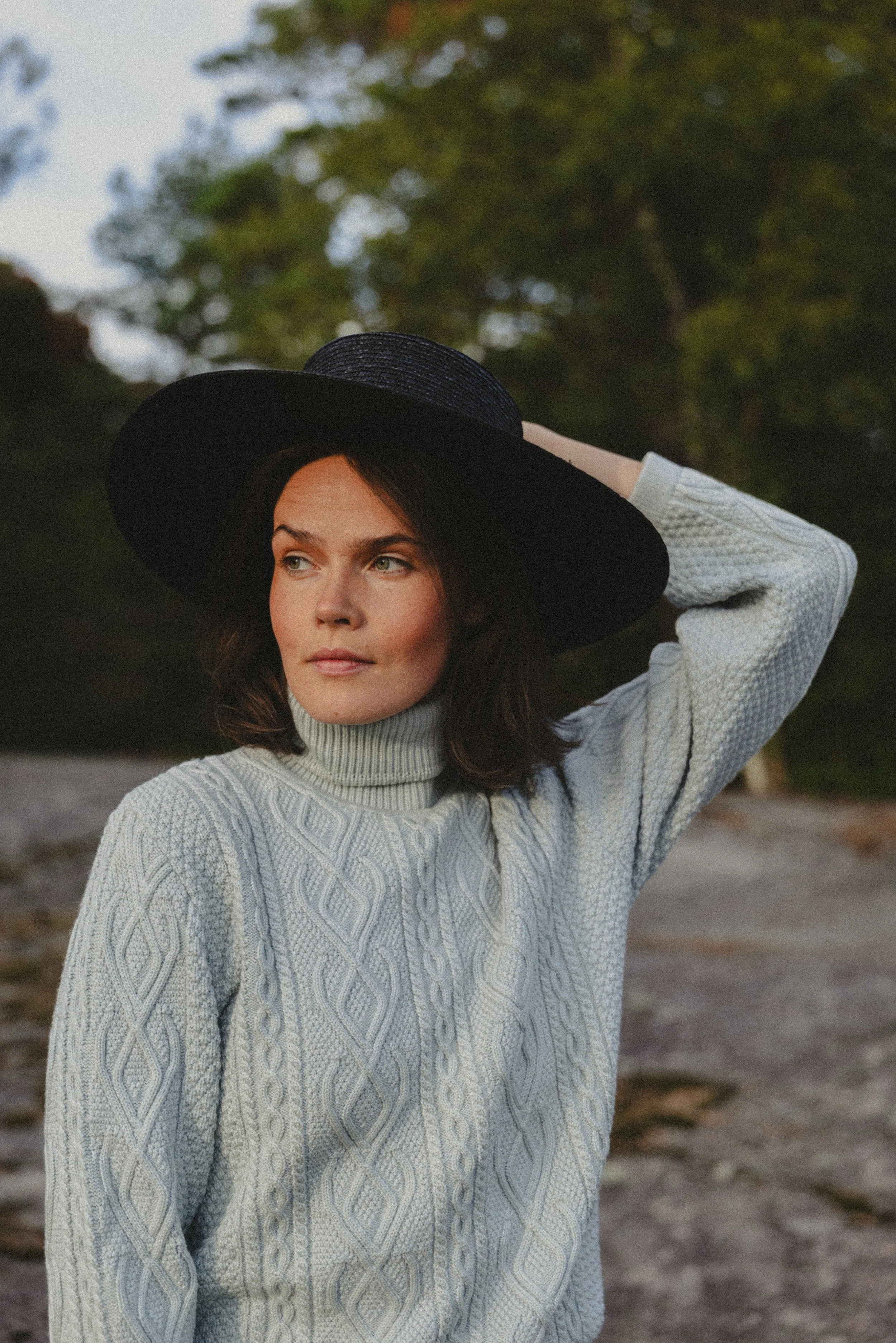 A woman in a gray cable-knit turtleneck sweater and a large black hat, outdoors with blurred green trees in the background.