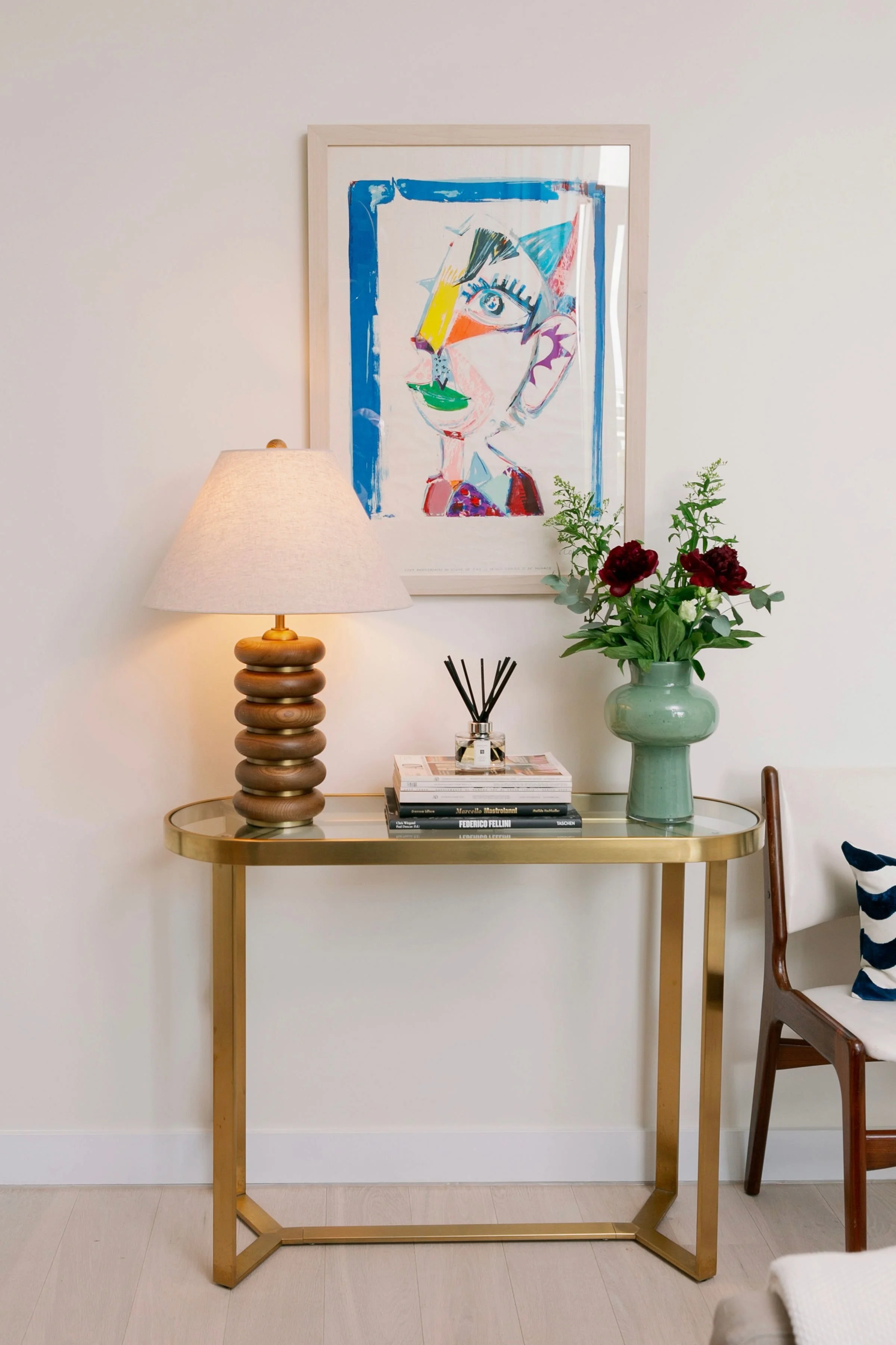 A gold-colored console table with a glass top, a lamp with a wooden base and beige shade, a vase with dark pink and white flowers, and a reed diffuser. A painting in a white frame featuring a colorful, abstract portrait of a woman is hanging on a plain white wall.