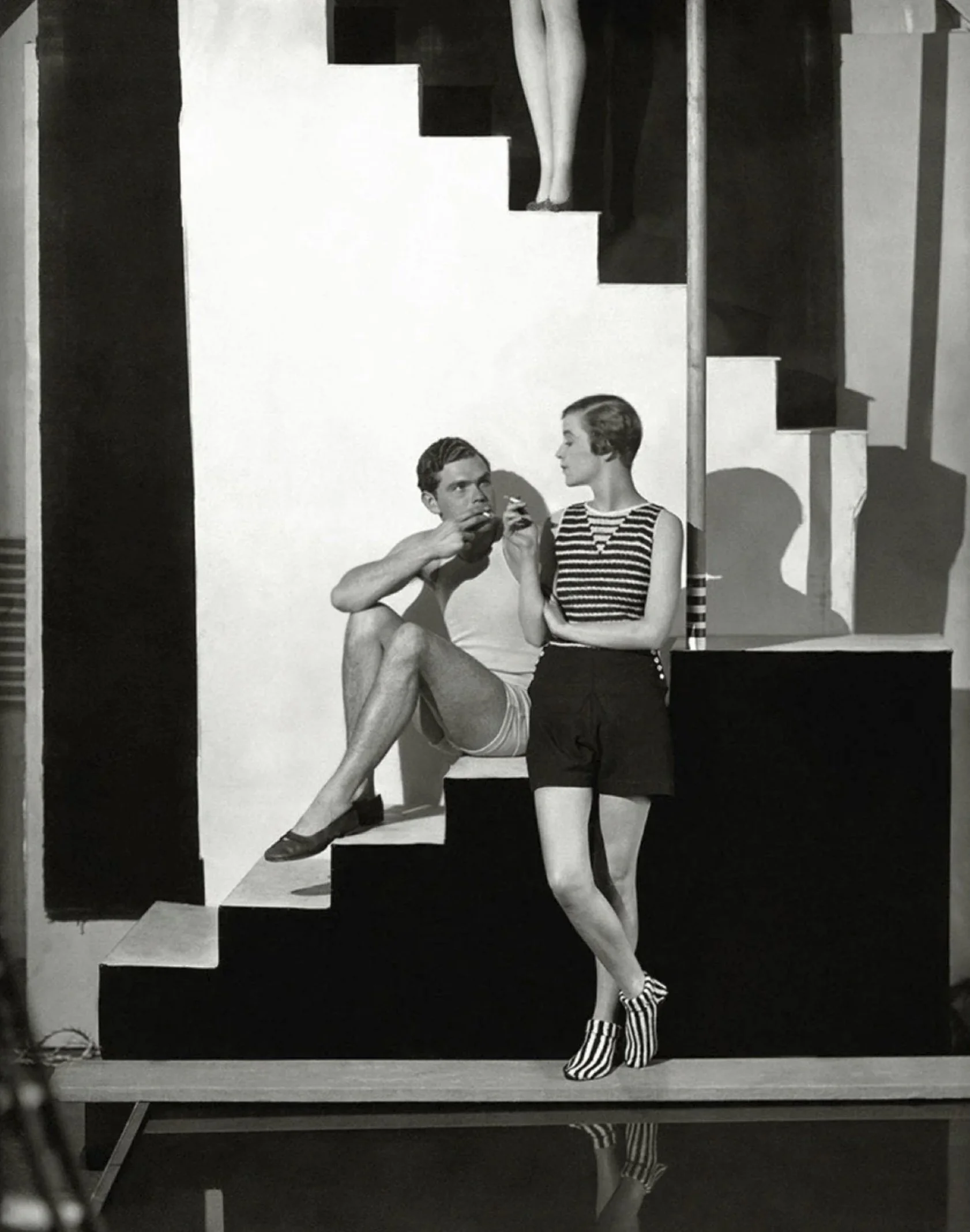 Black and white photograph of a man and a woman sitting on a staircase, with the man smoking and the woman looking at him, and a mannequin's legs visible behind them on the staircase.