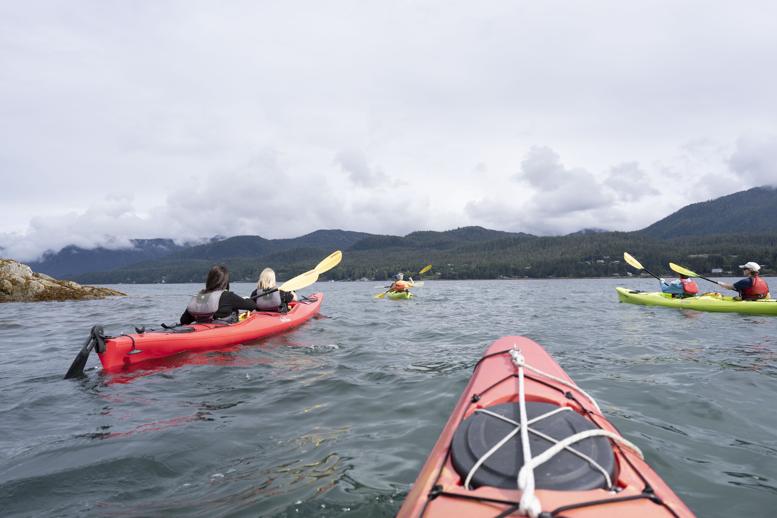 Looking out over the tip of a kayak at three more kayaks in water surrounded by forest.