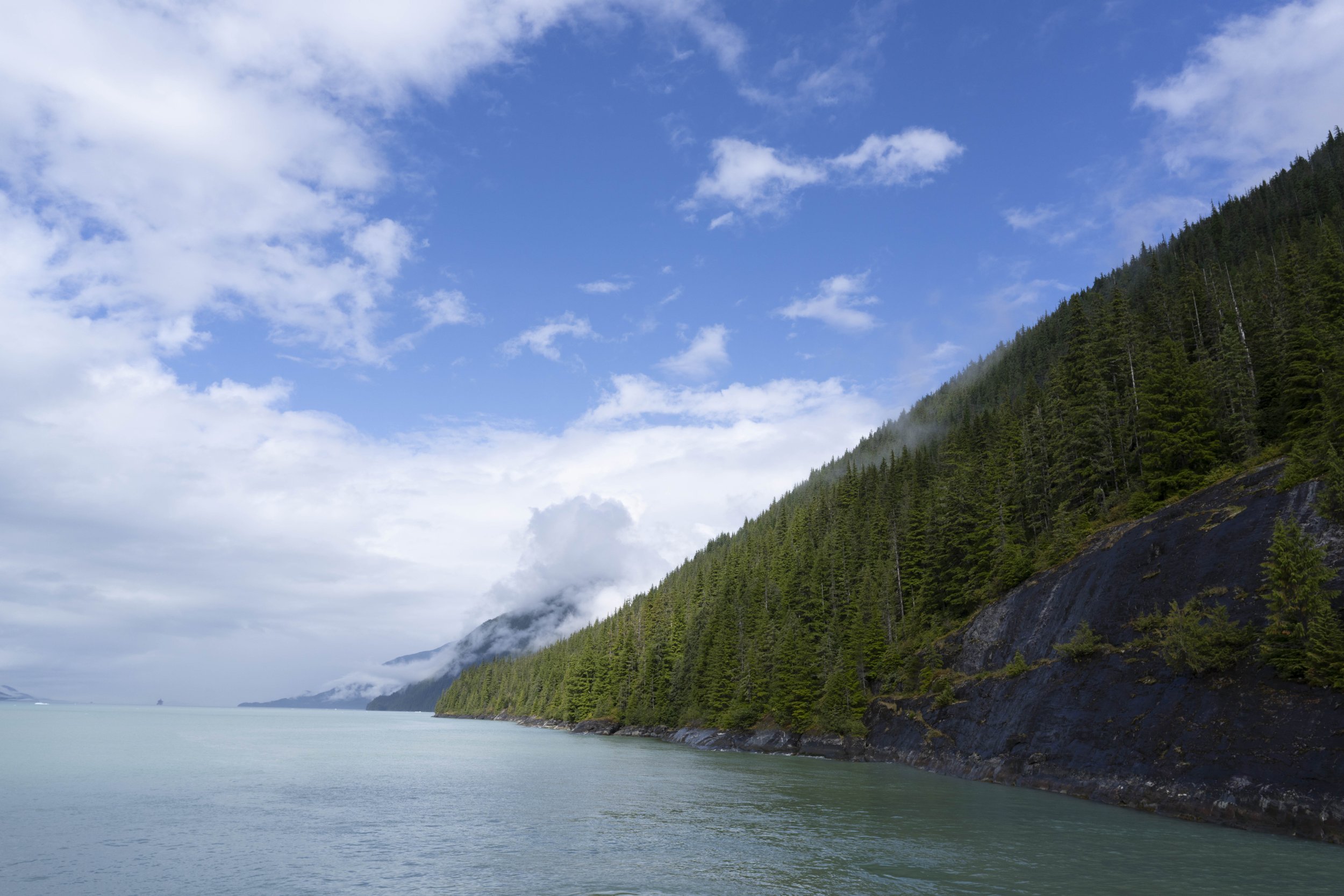 Varied clouds and blue sky over a forested mountain next to the water.