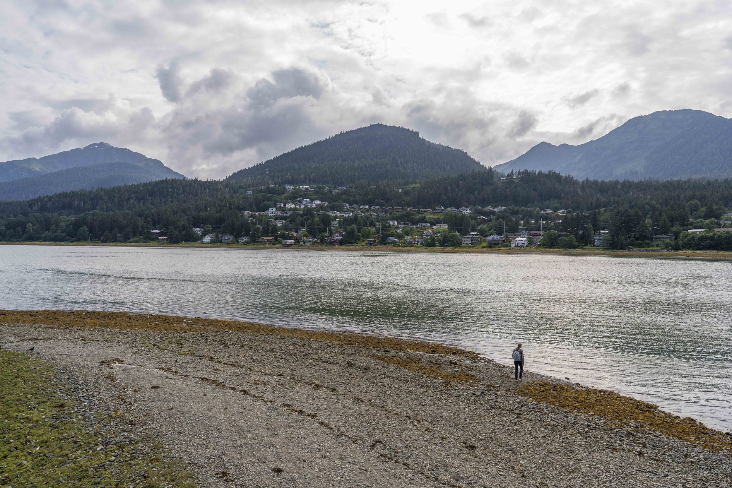 Nicole walking on a rocky beach with water, houses, and mountains in the background.