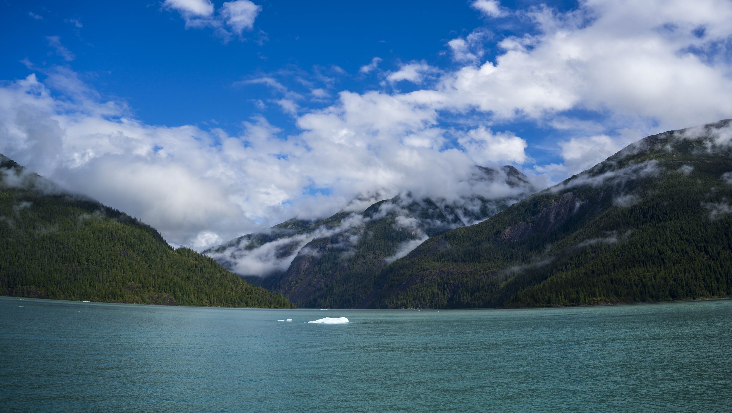 Low clouds over forested mountains with water and an iceberg in the foreground.