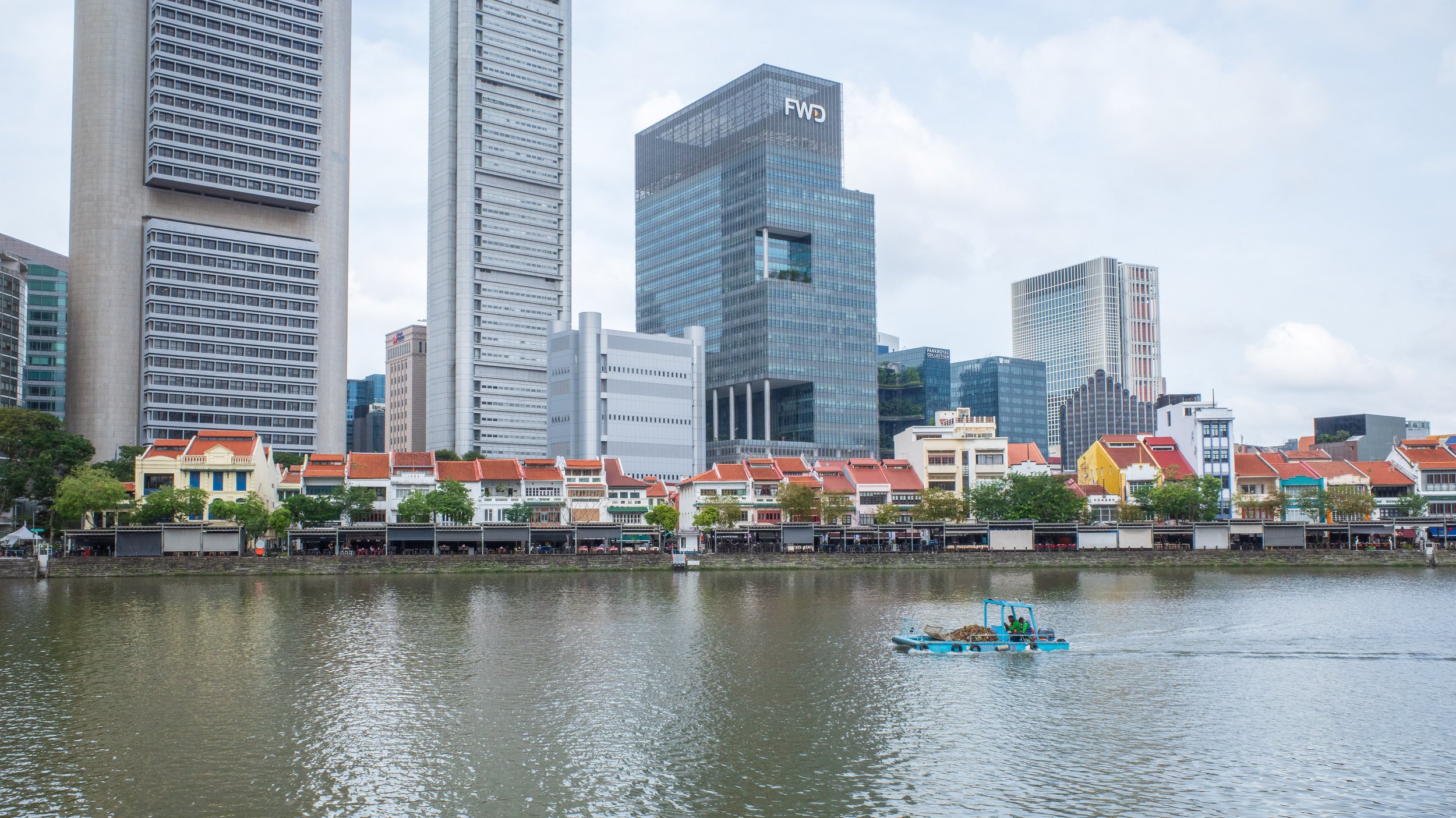 A small trash barge floating across the water in front of small buildings in front of skyscrapers.