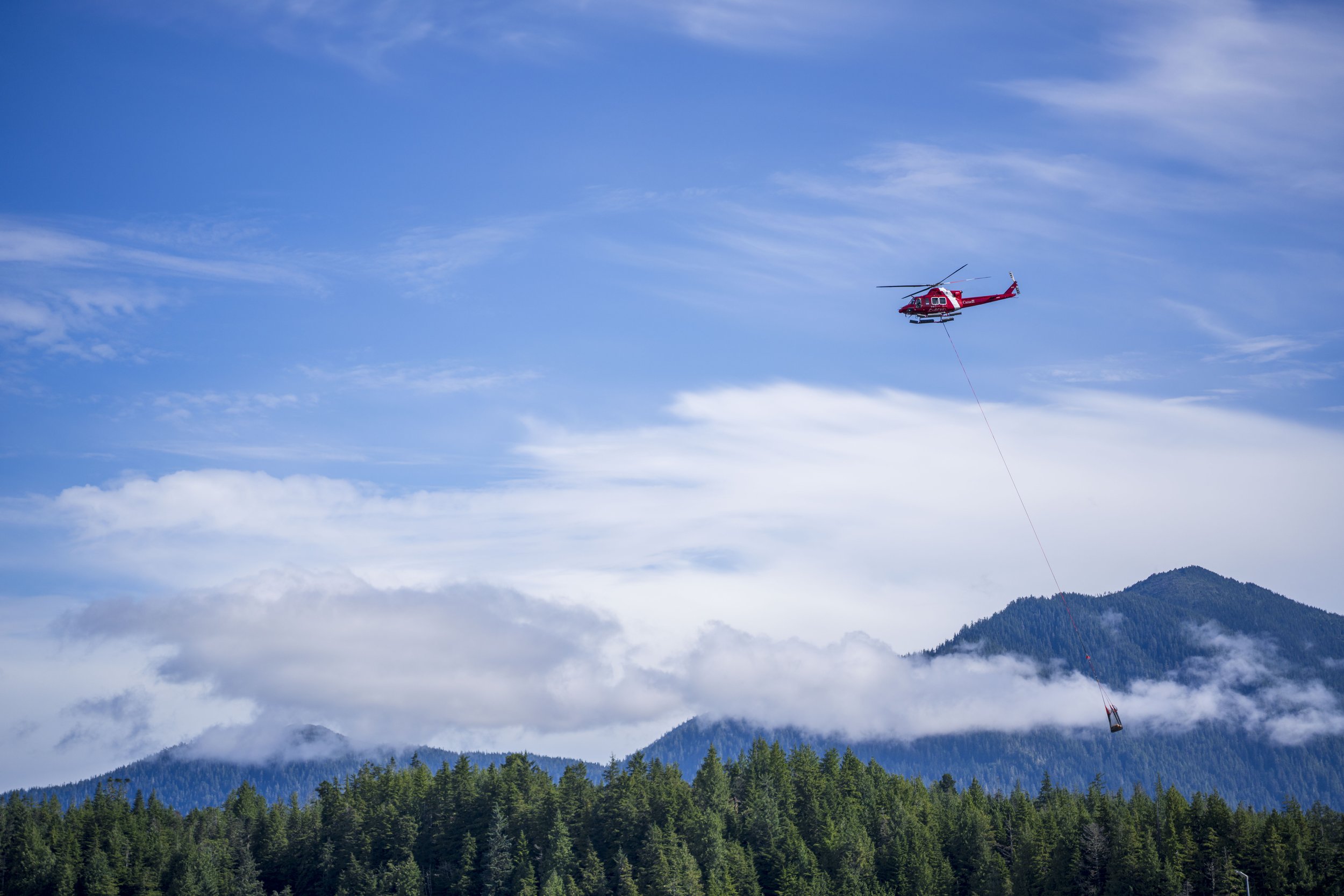 A helicopter with something hanging from the bottom flying over trees.