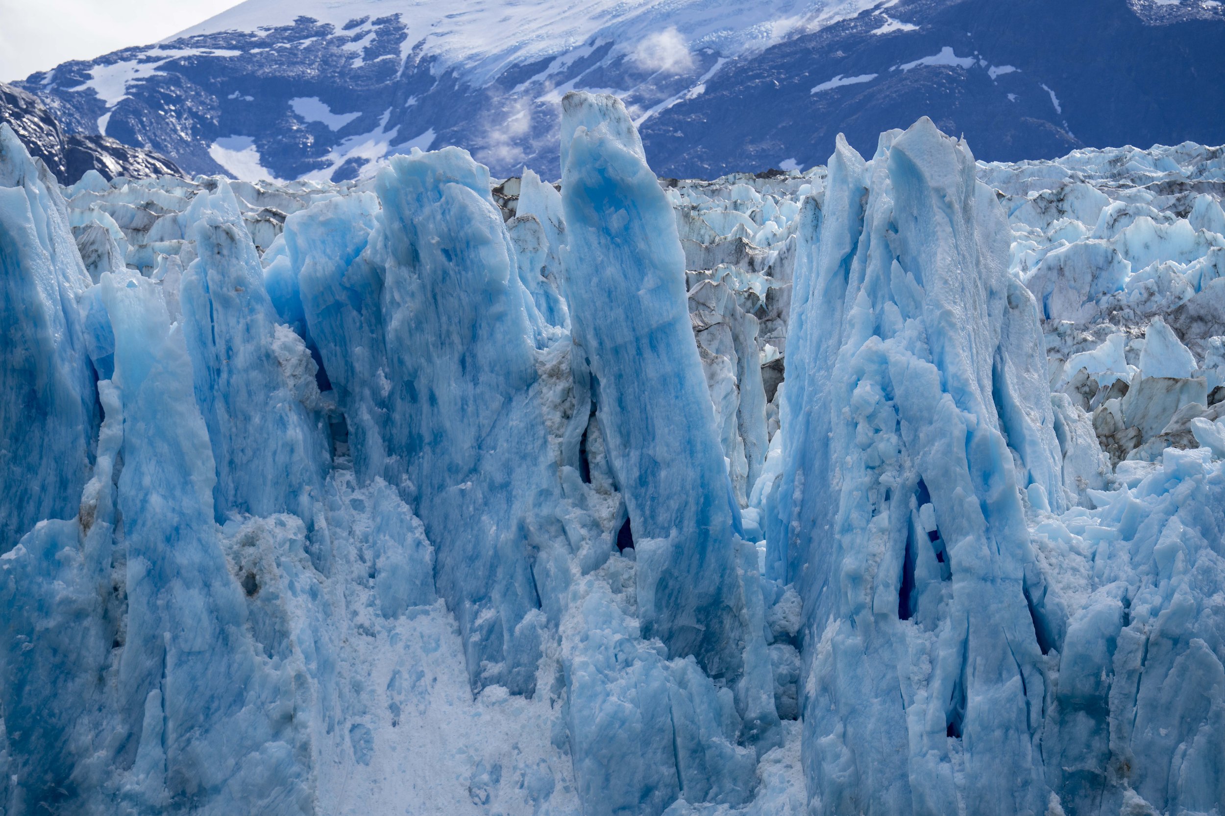 Spikes of bright blue glacier with snow-capped mountains in the background.