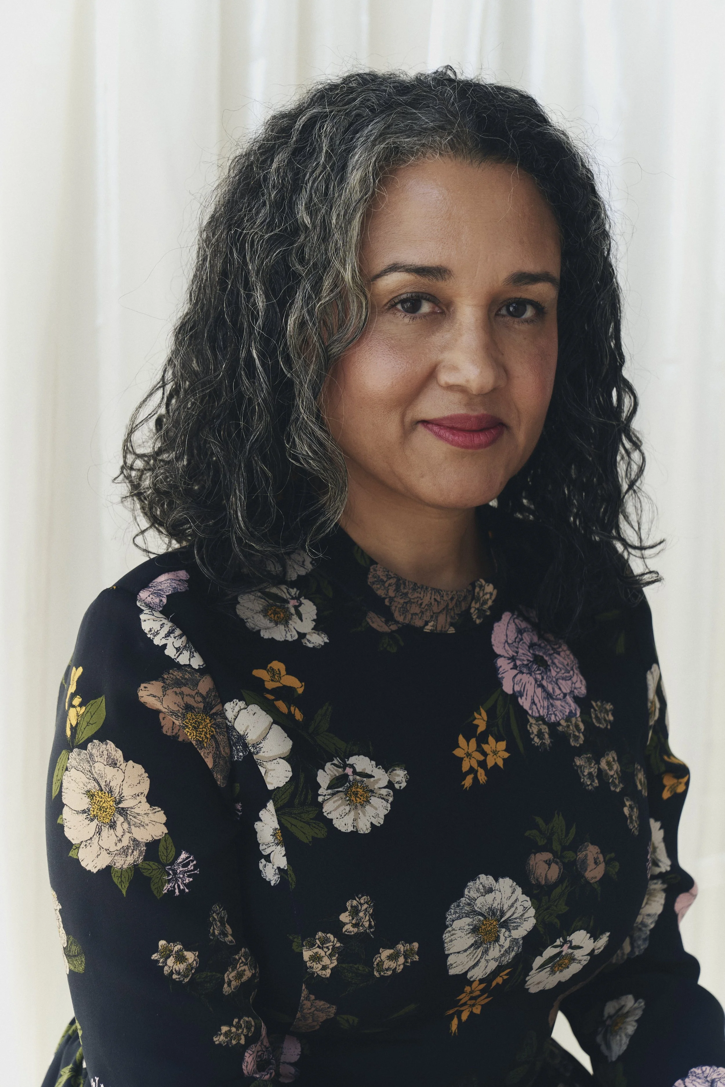 A woman with curly, salt-and-pepper hair wearing a black floral dress with pink, white, and orange flowers poses in front of a plain white curtain.