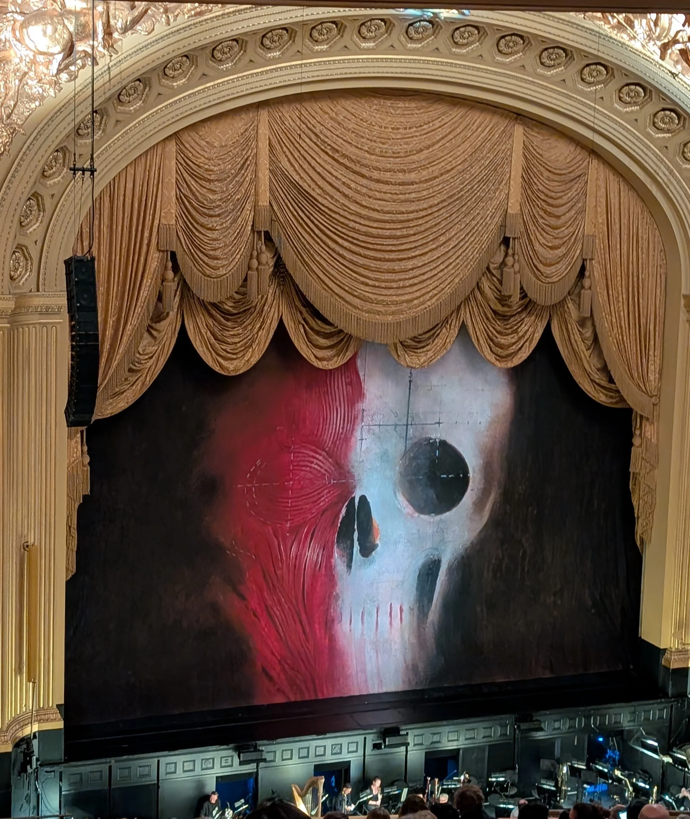 Orchestra pit below image of skull with blood red muscle across half of the skull projected onto the theater stage between gold curtains.