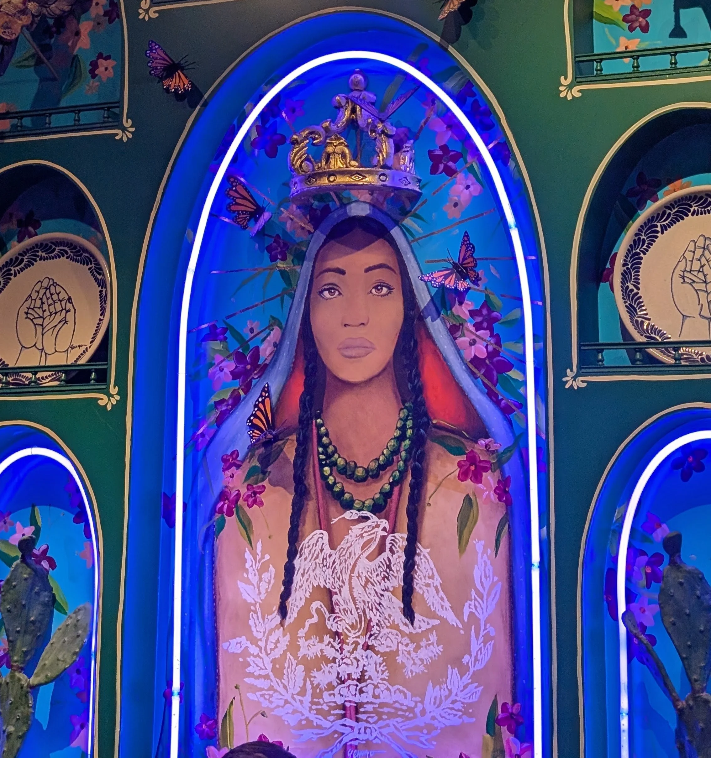 Religious icon art of woman with braids and cloak over her head with a crown floating above her and butterflies surrounding her.