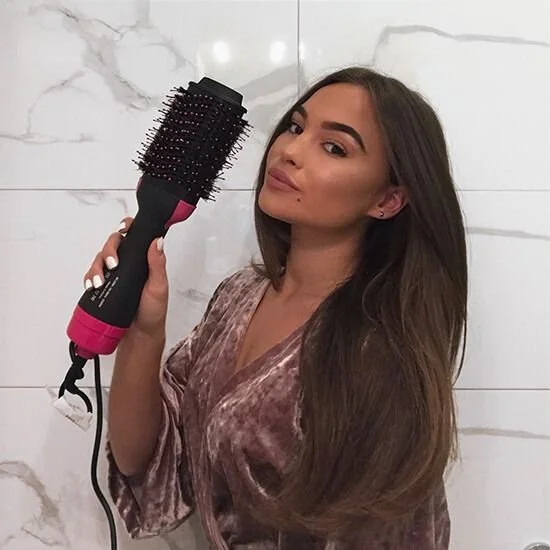 A woman with long brown hair holding a black and pink round hairbrush with a cord, standing against a white marble tiled wall.