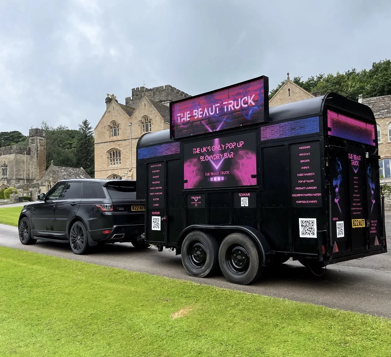 Black mobile promotional trailer for The Beauty Truck, showcasing pink and purple neon graphics and text, parked on a street with a black Range Rover nearby, with historic stone buildings and greenery in the background.