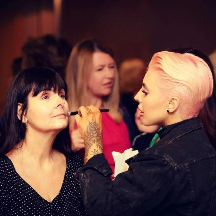 A woman with dark hair and a polka dot top having her makeup or face painted by a person with short pink hair and a tattooed arm, at an indoor event.