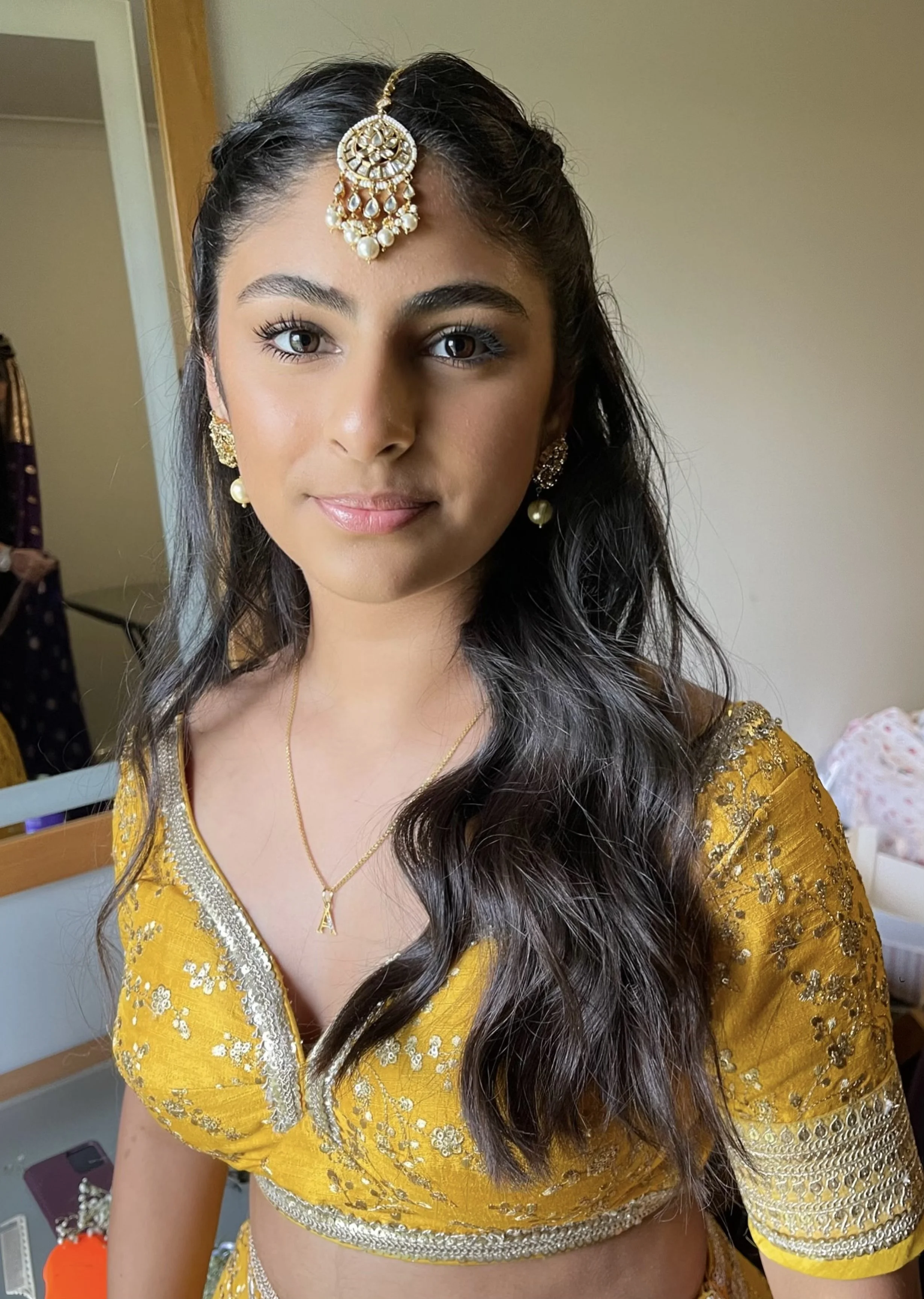 A woman wearing traditional Indian attire with gold embroidered yellow blouse, jewelry including earrings, a necklace with a letter 'A' pendant, and a decorative headpiece, smiling at the camera.