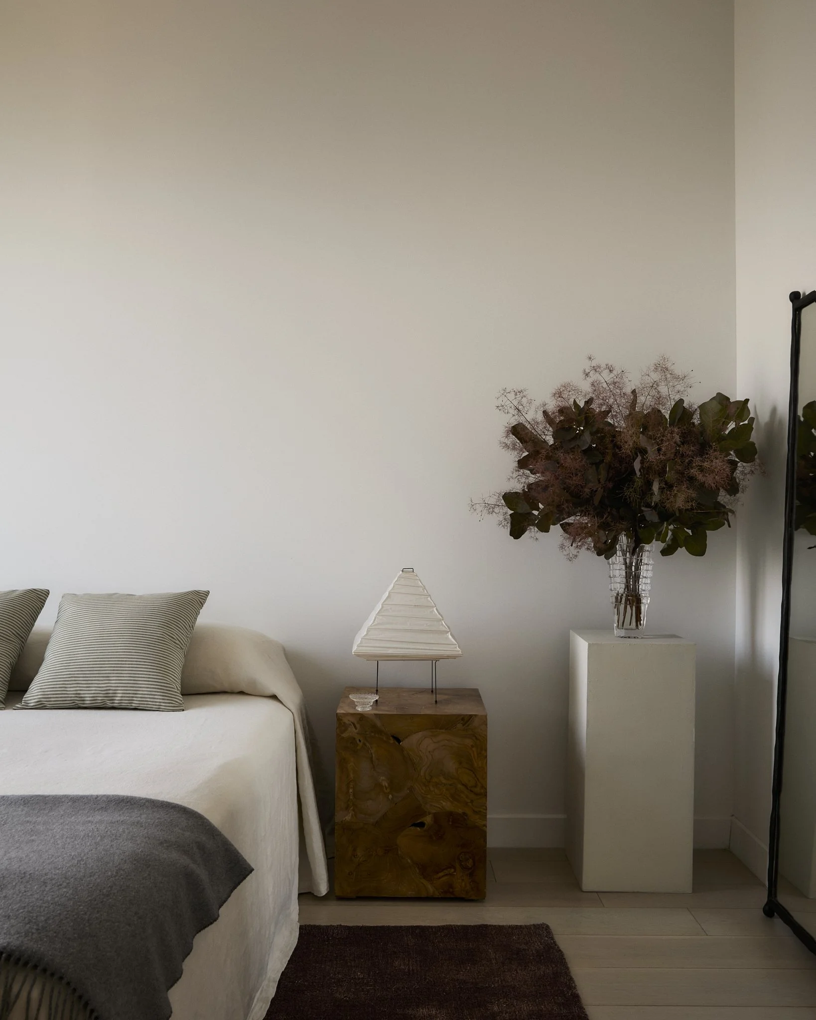Prospect Heights primary bedroom detail by Hallie Rose Design featuring a burl-wood nightstand, Japanese paper lantern lamp, striped pillows, and smoke-bush branches displayed on a plaster pedestal.