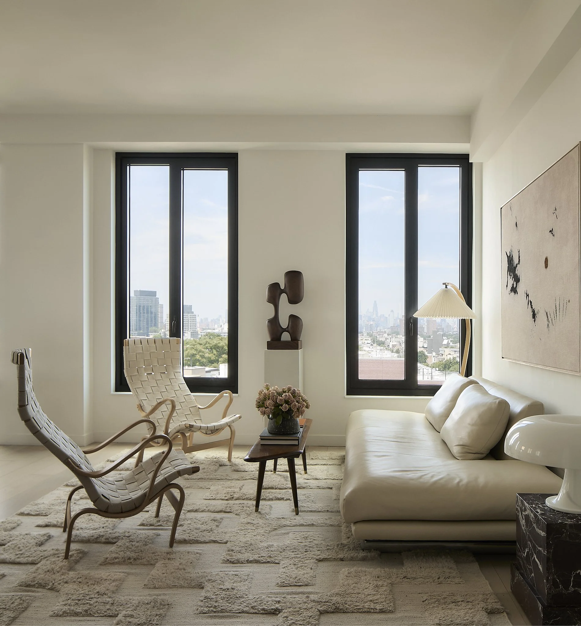 Modern living room with large windows showing city skyline, white leather sofa, unique wooden sculpture, beige area rug, woven chaise lounge, floor lamp, and black marble side table with a white vase.