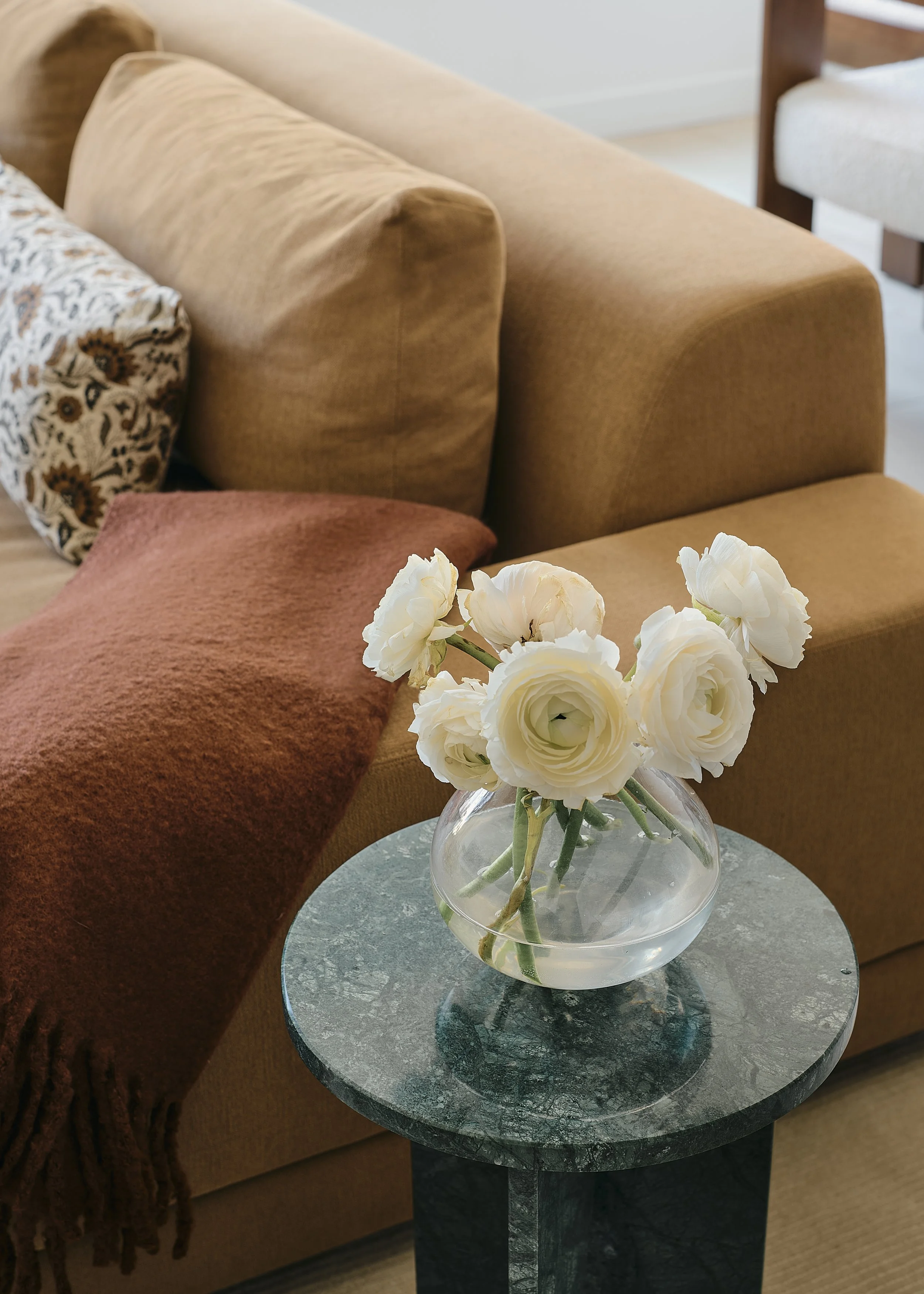 Prospect Heights living room detail by Hallie Rose Design featuring a green marble side table, white ranunculus in a glass vase, camel-toned sofa, rust wool throw, and patterned textile pillow.