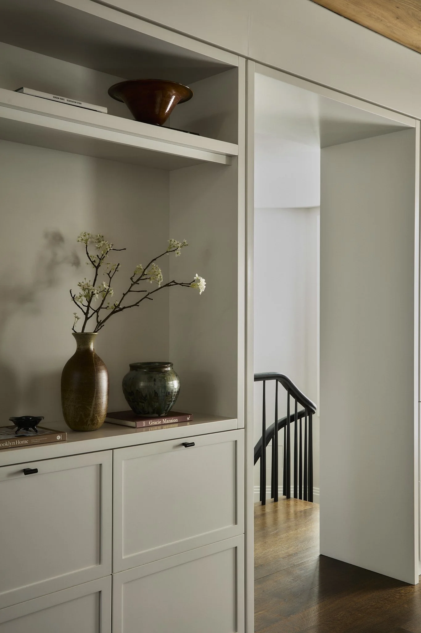 A white built-in cabinet with decorative vases and books on top, a small branch with white flowers in a vase, and a black staircase railing nearby. The floor is wooden, and there is a doorway leading to a hallway.