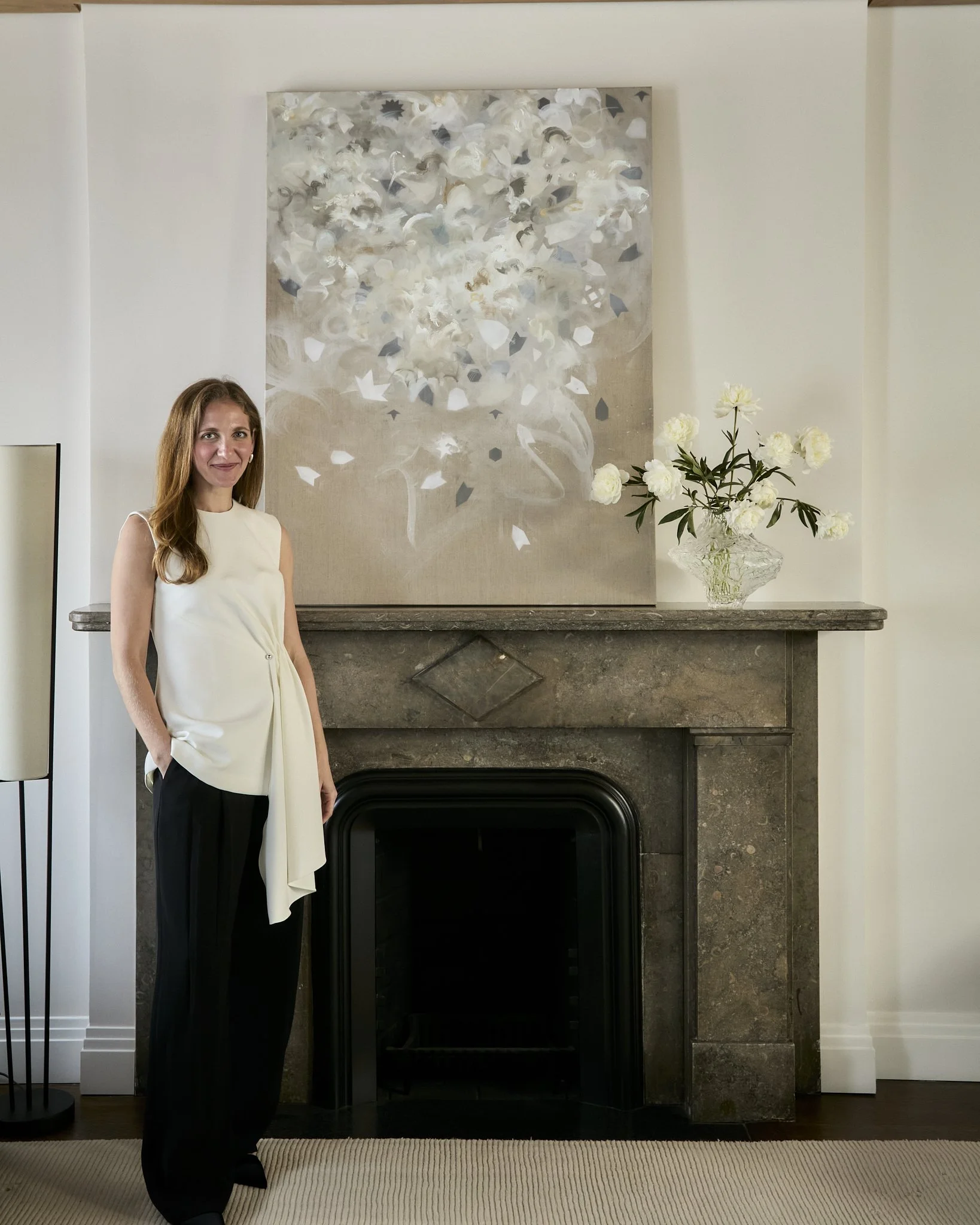 Interior designer Hallie Rose in a Brooklyn Heights residence beside abstract artwork, a stone fireplace, and white peony floral arrangement.